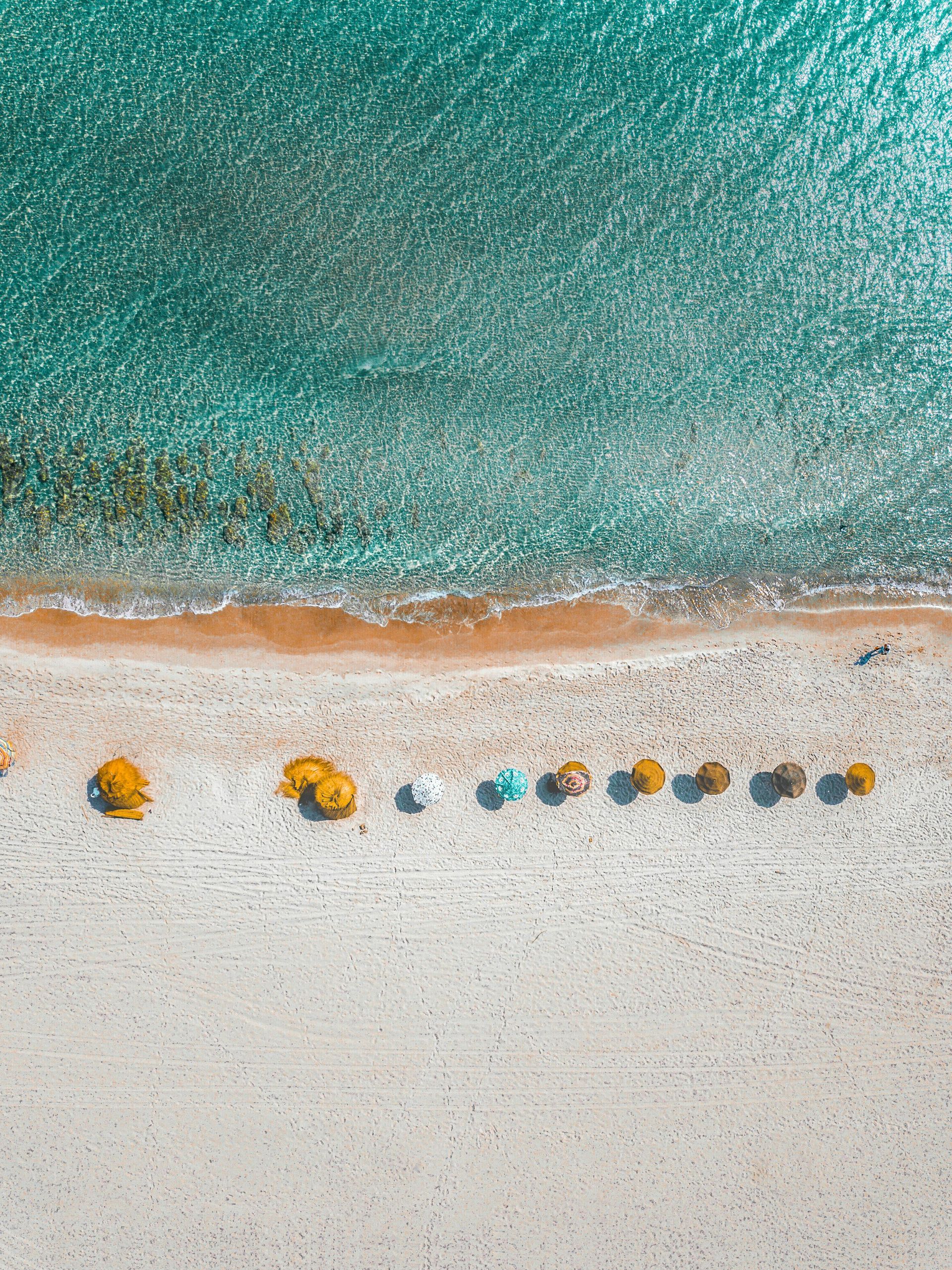 An aerial view of a white sand beach with a row of colorful umbrellas along the turquoise shoreline.