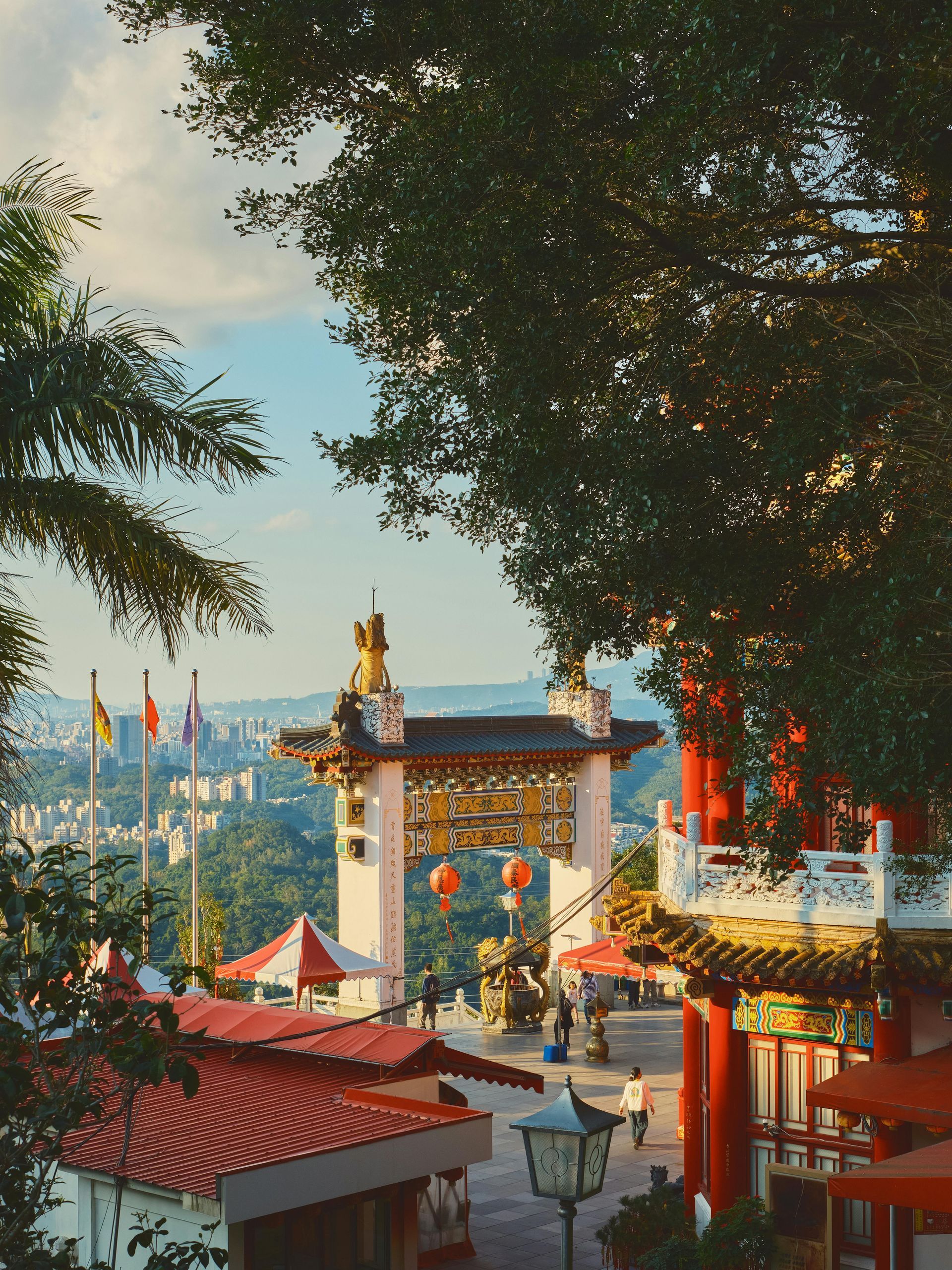 A view of a traditional Chinese-style archway and temple buildings set against a lush, hilly landscape at sunset.