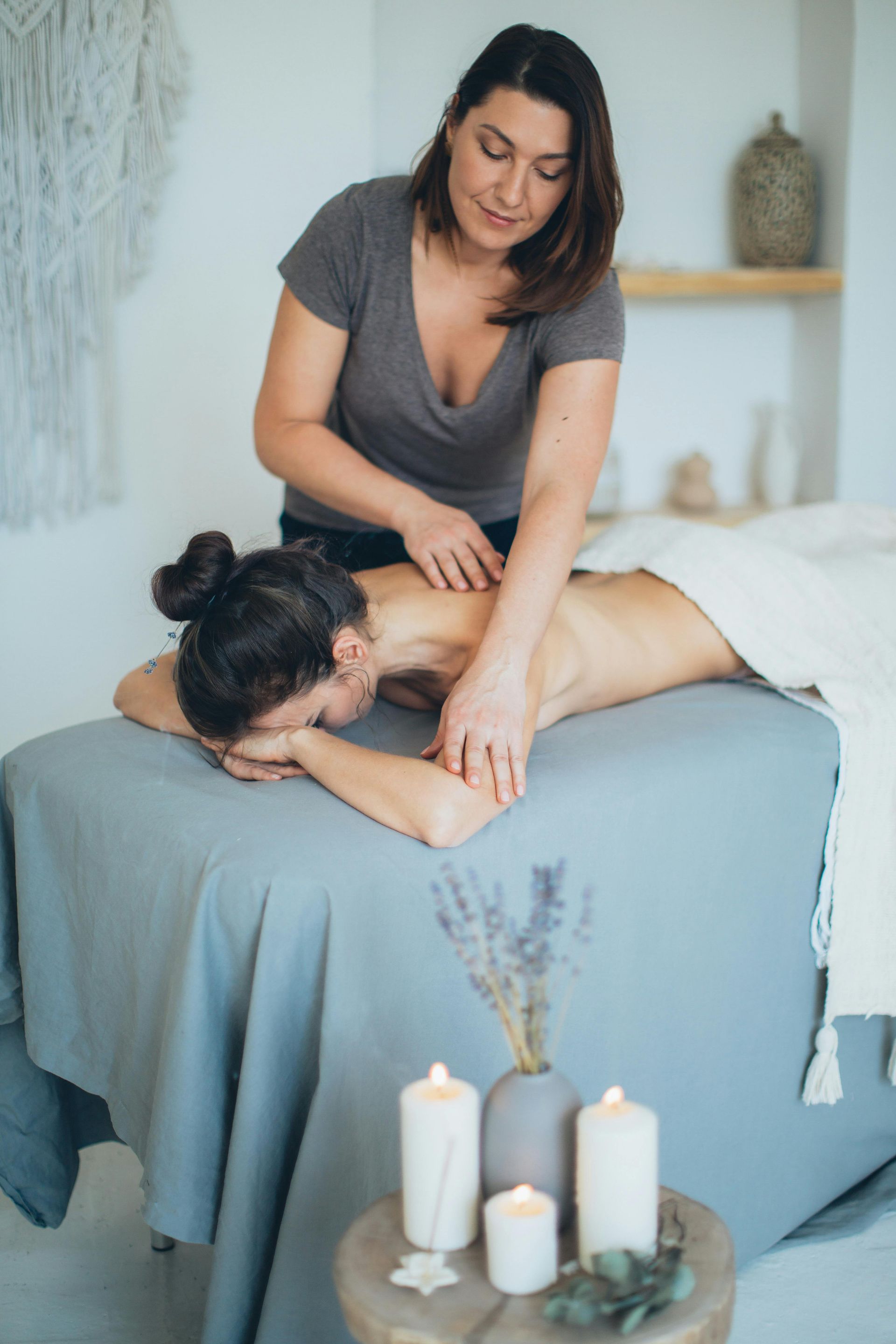 A massage therapist in a grey t-shirt massages the back of a client on a treatment table, with candles in the foreground.