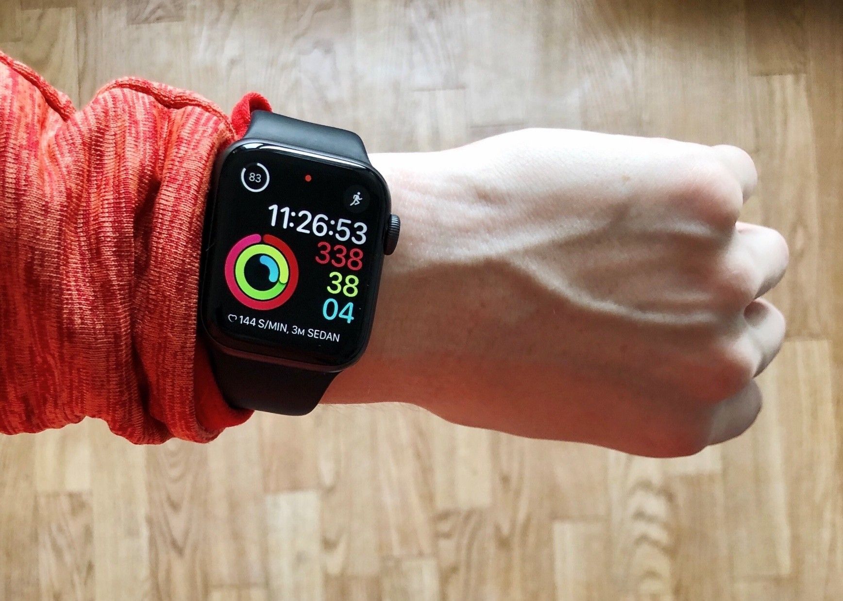 Wrist wearing a black smartwatch showing activity rings on a hand over a wood floor
