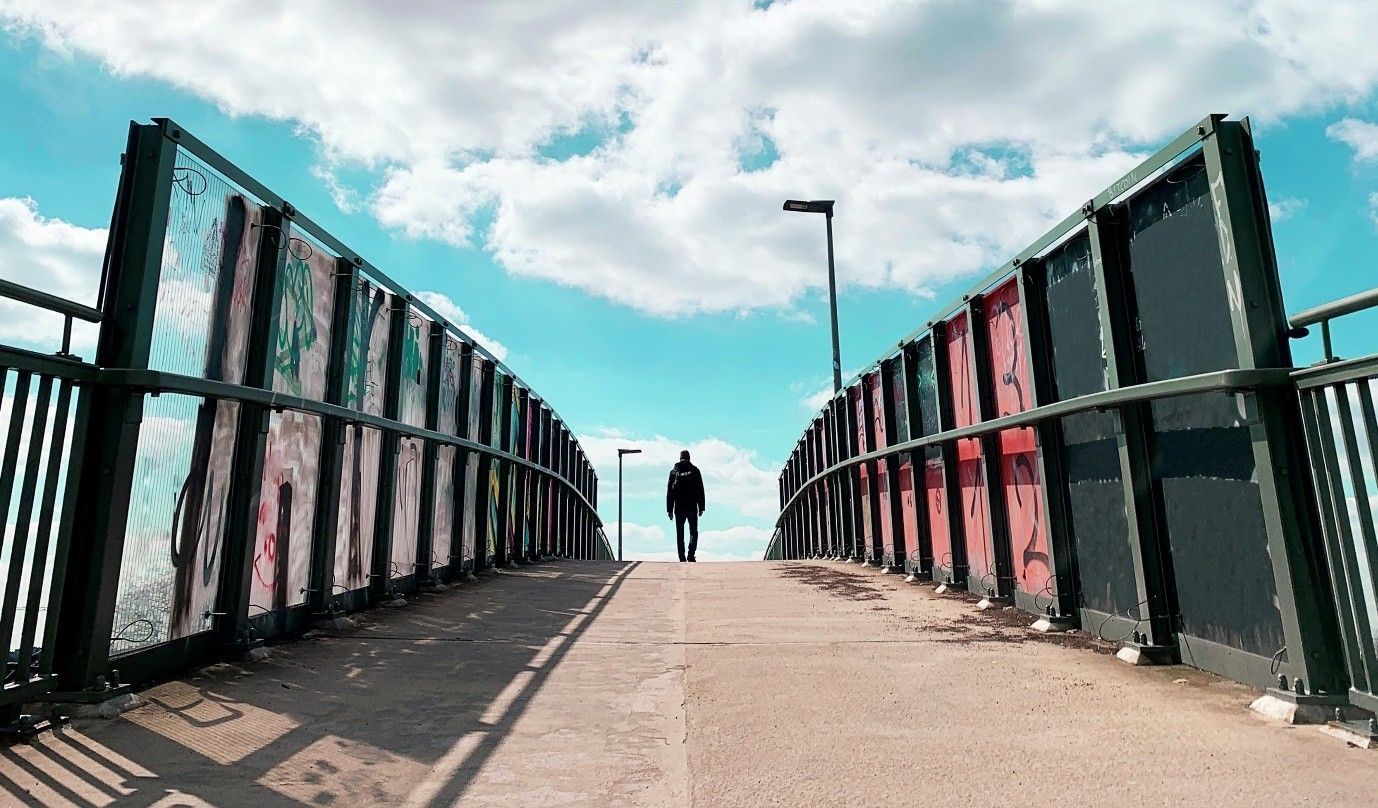 Person walking on a pedestrian bridge with green railings under a bright cloudy sky