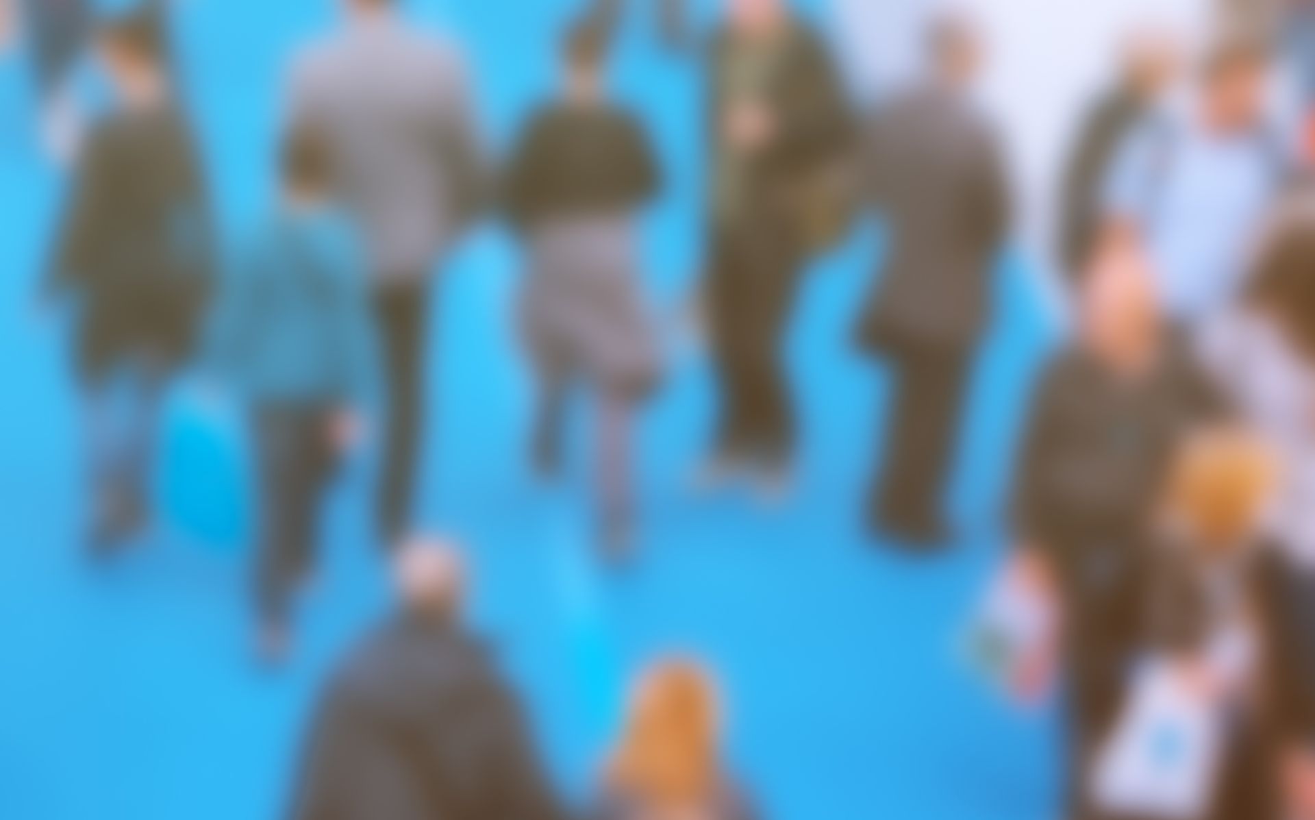 Blurred crowd of people standing and walking on a bright blue floor at an event.