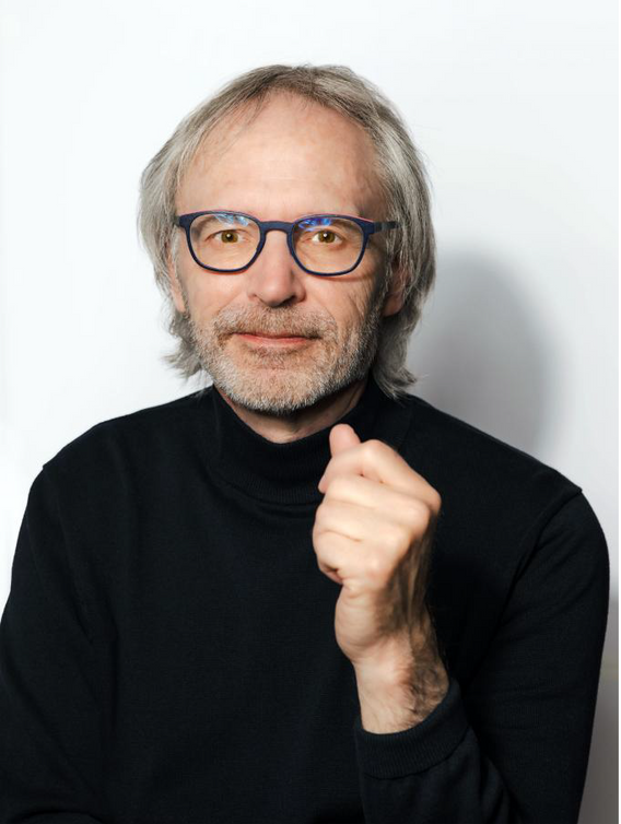 Portrait of Tom Lawry with glasses and gray hair, wearing a black turtleneck against a plain light background.