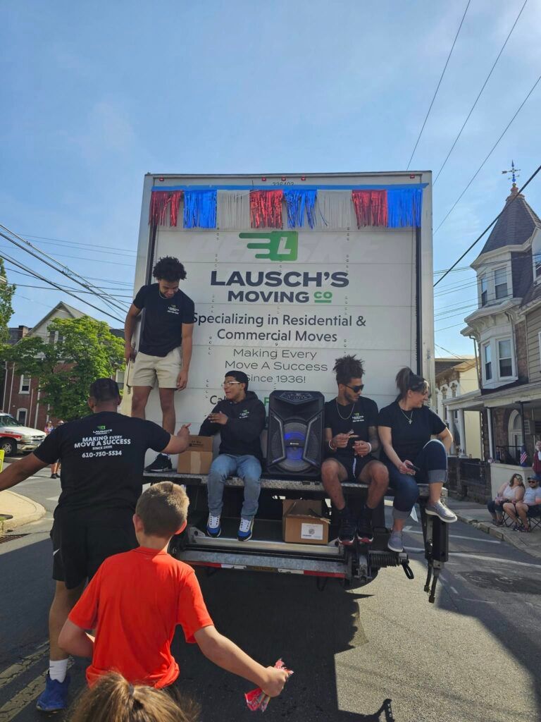 People in matching black uniforms on a Lausch's Moving truck during an outdoor parade on a sunny day.