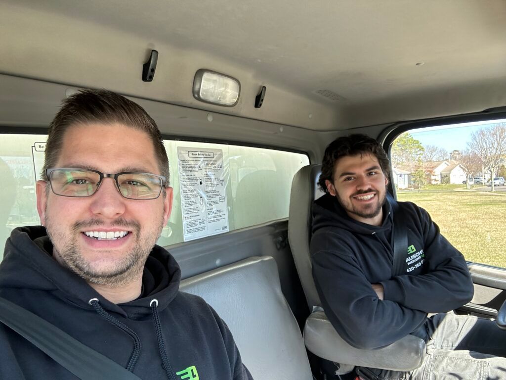 Two people smiling inside a vehicle cab, wearing dark hoodies and seatbelts, with a document taped to the window.