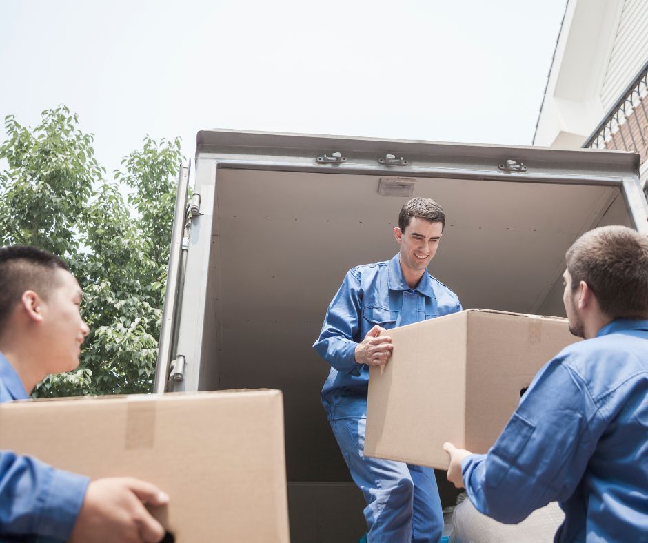 Three movers in blue uniforms work together to load or unload cardboard boxes from the back of a moving truck.