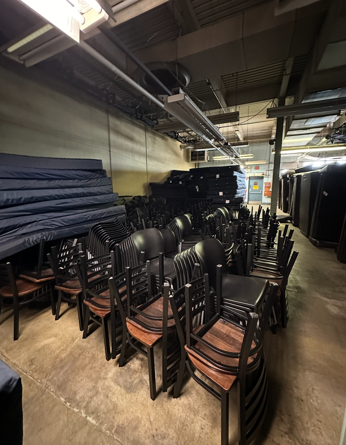 Rows of dark brown, stacked chairs in a dimly lit industrial storage area with stacks of blue mattresses against the wall.
