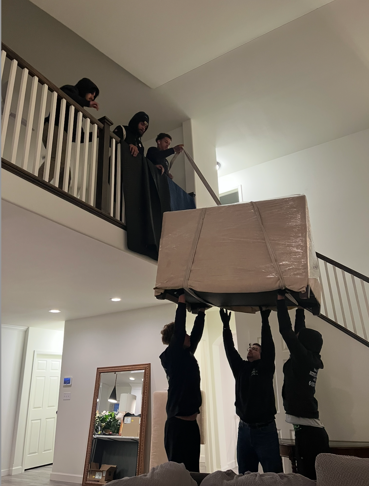 Three people below lift a large, wrapped rectangular object while three others watch from an upstairs hallway railing.