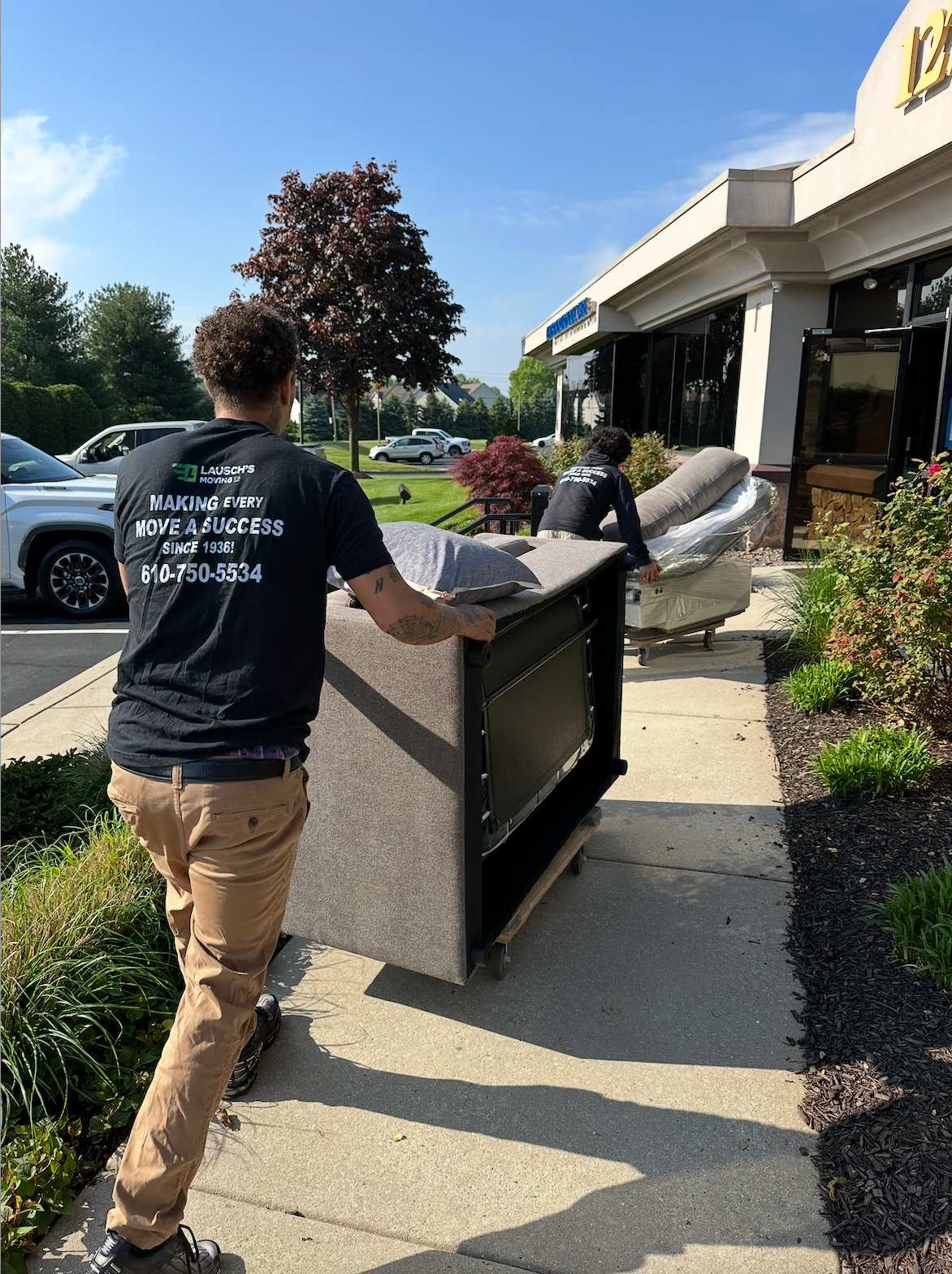 Two movers transport large furniture on dollies along a sidewalk outside a retail storefront.