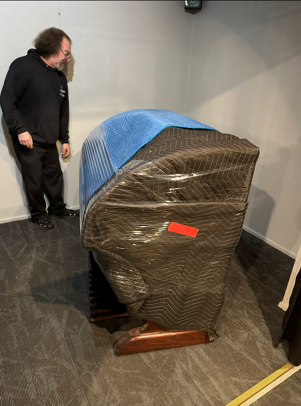 A person standing beside a large piano wrapped in protective plastic and a blue blanket in a room with gray carpet.
