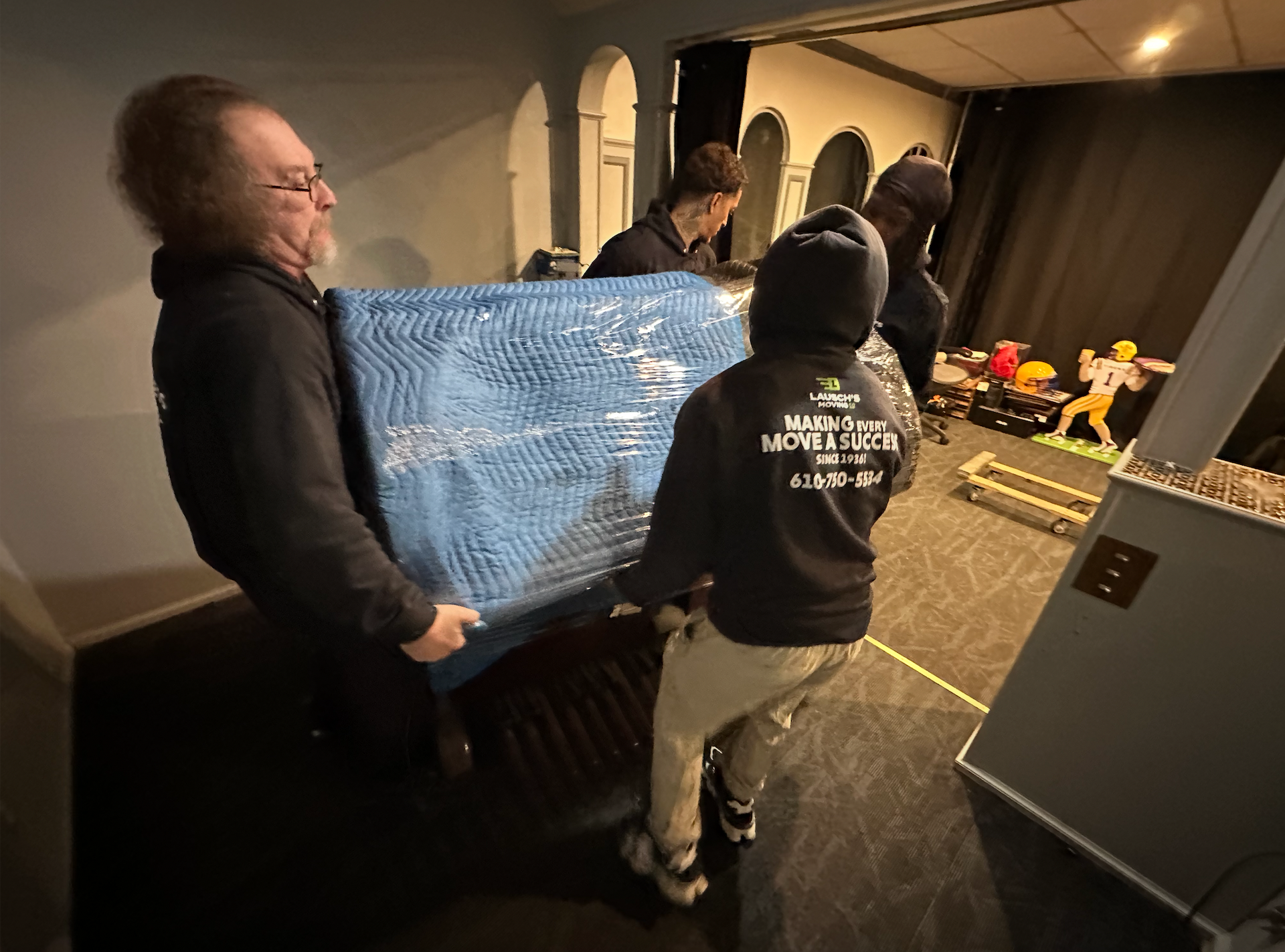 Movers in dark hoodies carry a large, blue-wrapped item through an indoor space.