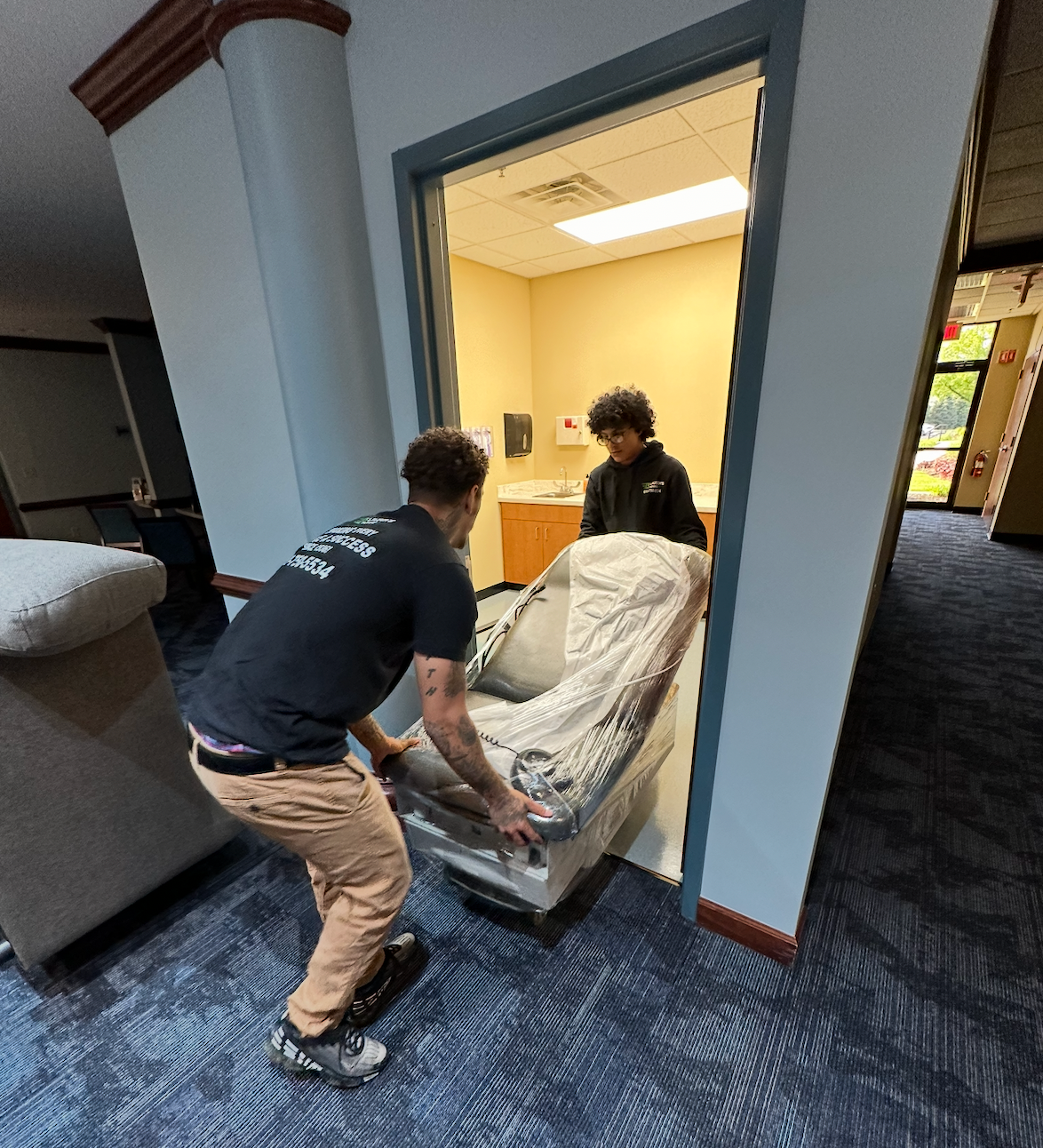 Two movers in work attire maneuver a wrapped piece of furniture through an office doorway.