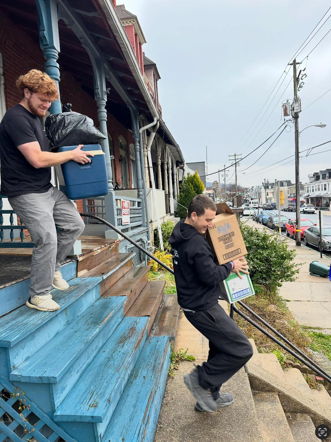 Two people carry boxes and bags down the steps of a porch toward a street.
