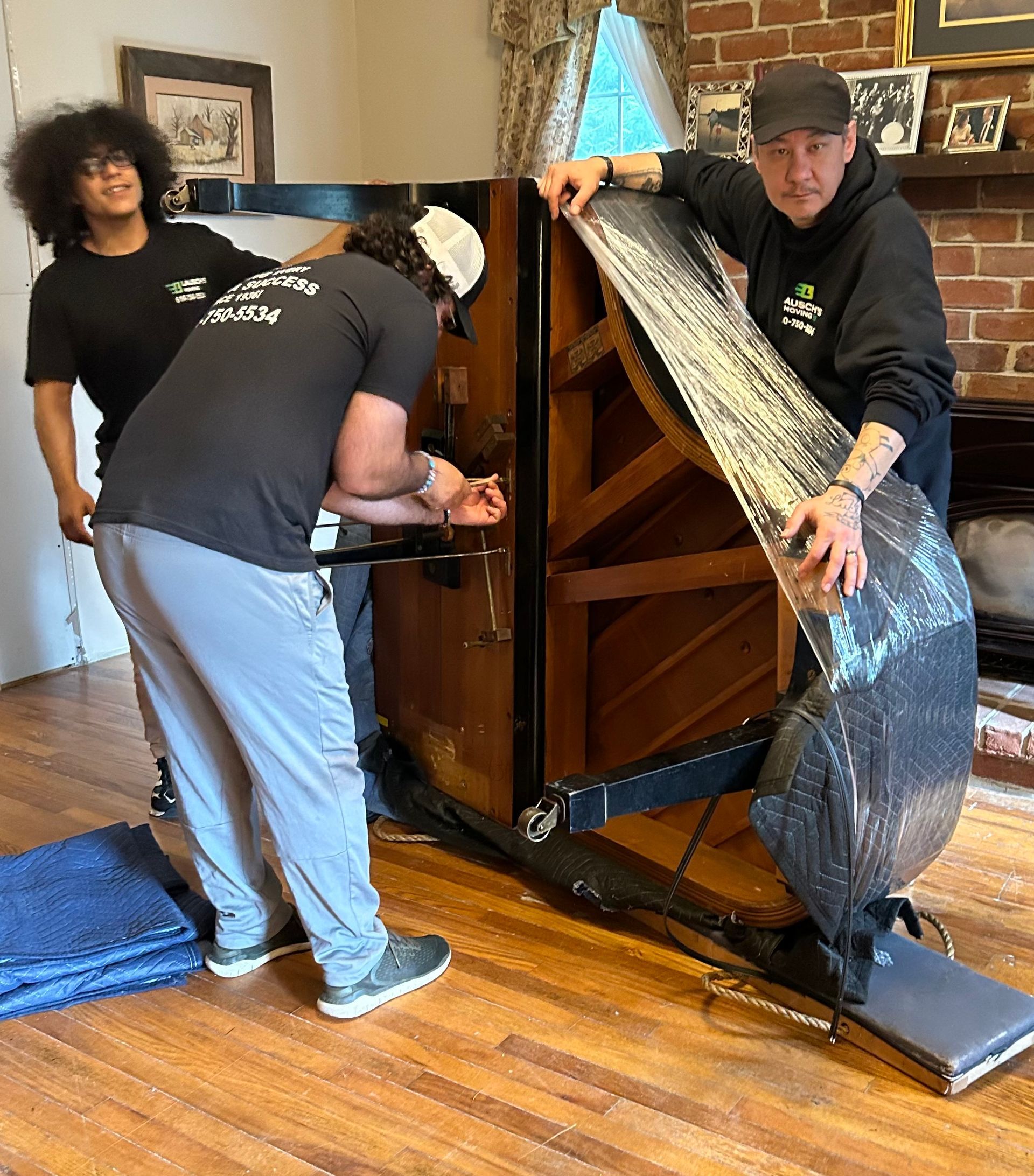 Three people work together to move and wrap a large, upright piano in a residential room with hardwood floors.