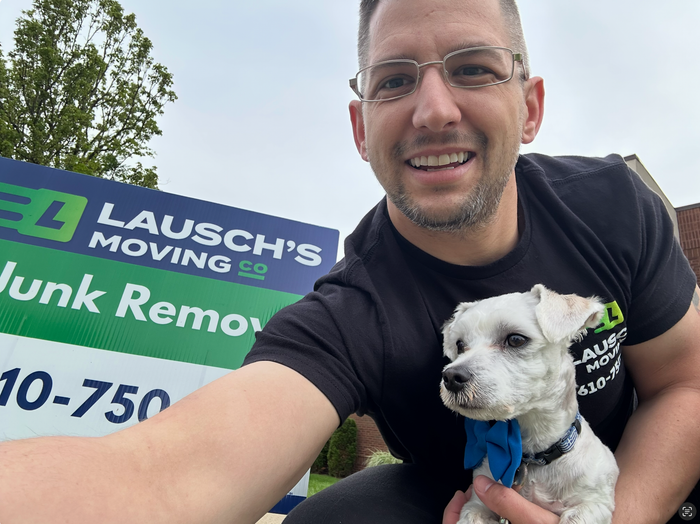 A smiling person holds a small white dog with a blue bow in front of a blue and green Lausch's Moving sign.