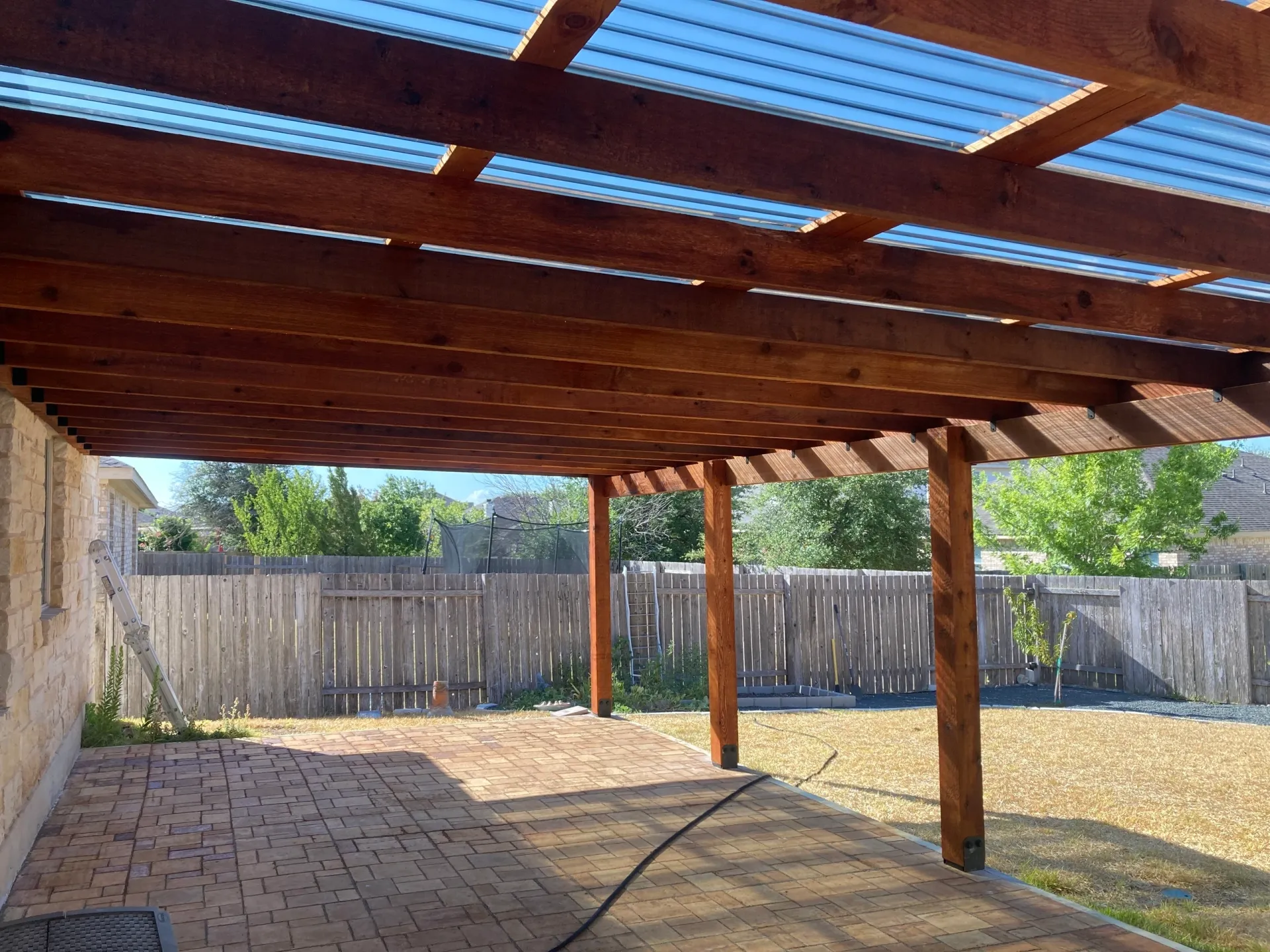 Wooden pergola patio over a brick and gravel backyard with a fenced perimeter and trees beyond