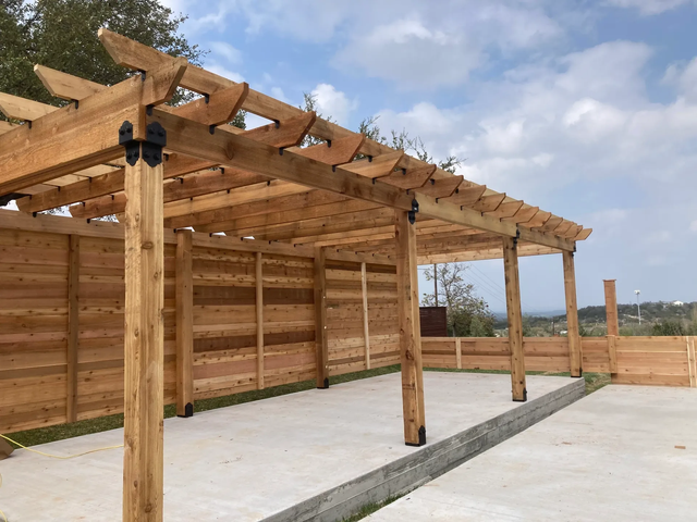 Wooden pergola framing a patio under construction on a concrete slab, with clear blue sky overhead