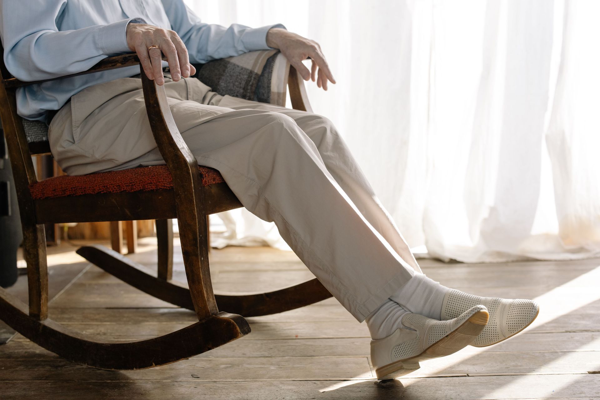 Person reclining in a wooden rocking chair by a sunlit window, legs stretched out on a rug.
