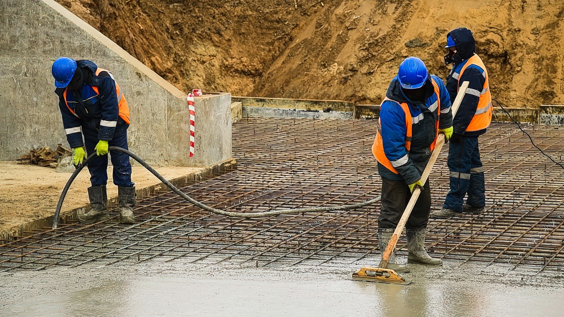 Bauarbeiter gießen Beton. Sie tragen blaue Helme, orangefarbene Westen und Arbeitsstiefel. Beton wird auf Bewehrungsstahl gegossen.