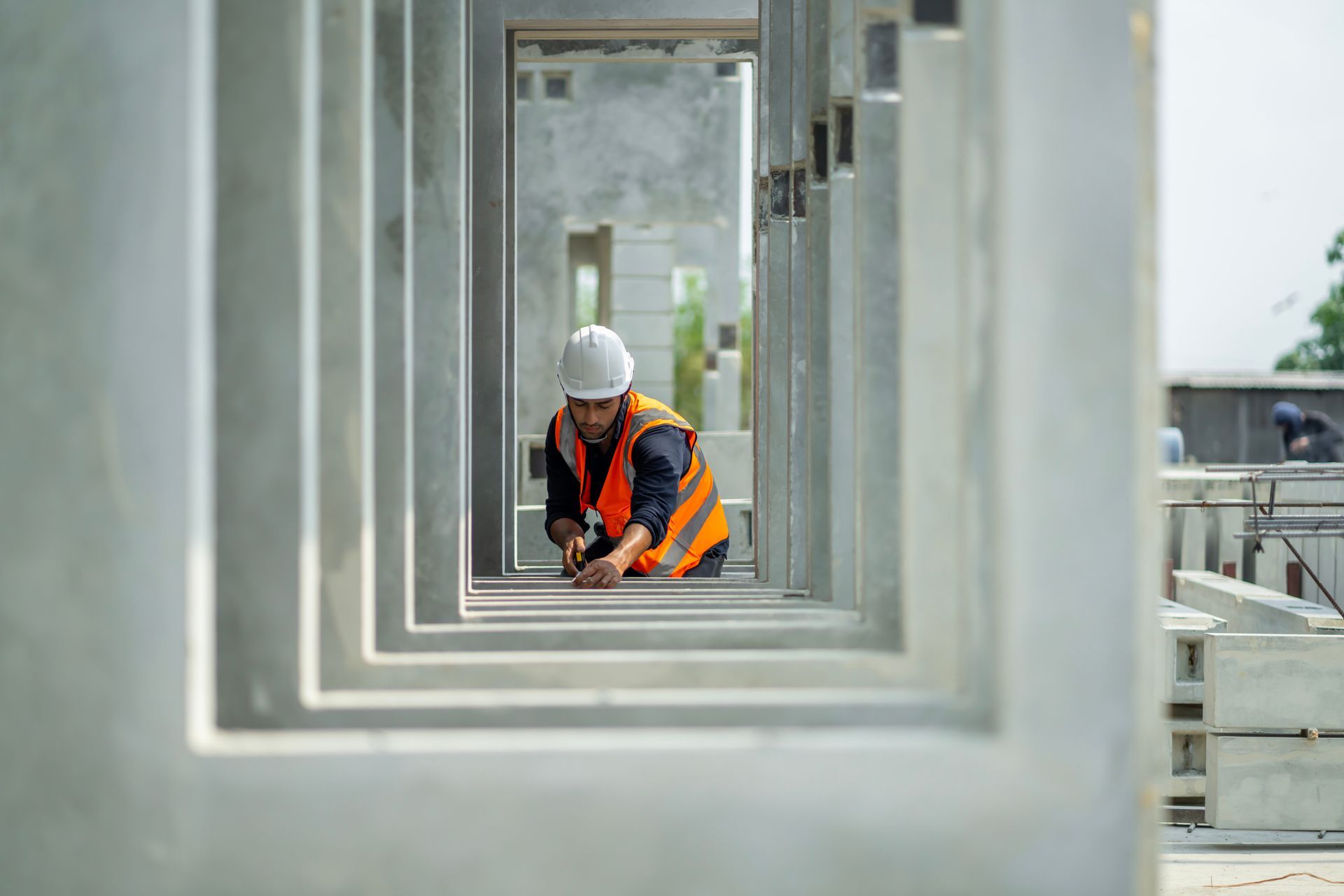 Bauarbeiter mit Schutzhelm und Weste misst auf einer Baustelle einen Betonrahmen.