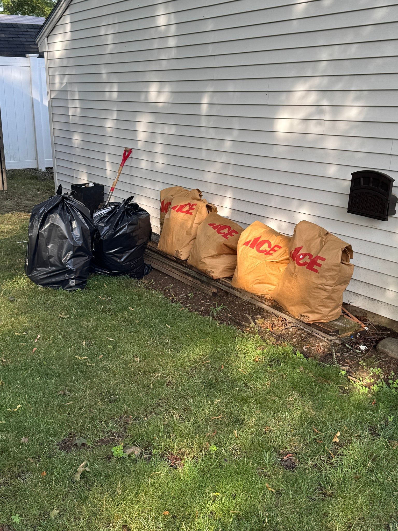 Trash bags and leaf bags lined up beside a house’s siding along the lawn.