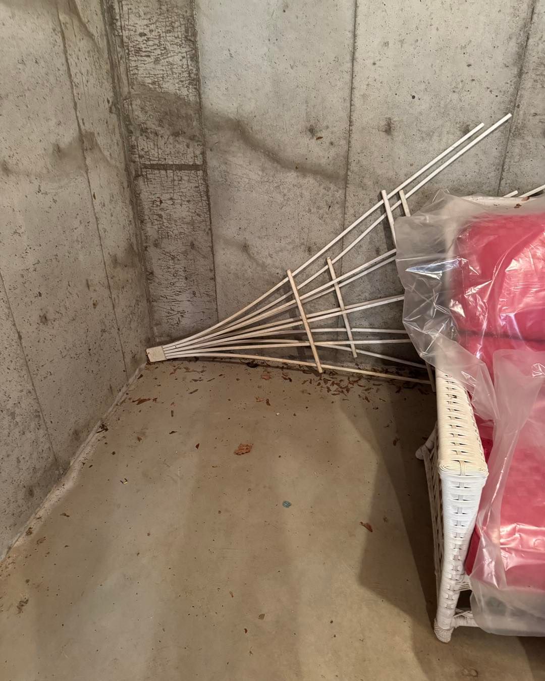 Collapsed white drying rack leaning in a concrete corner beside a pink-covered chair