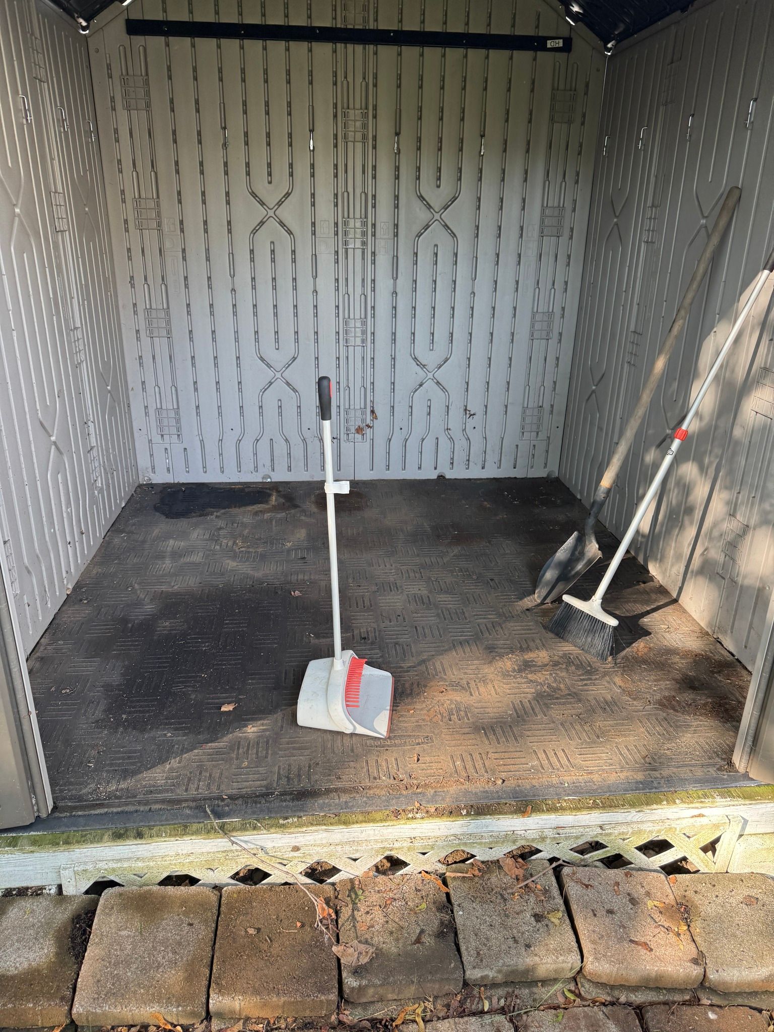 Small storage shed interior with two brooms, a dustpan, and patterned walls over a brick threshold