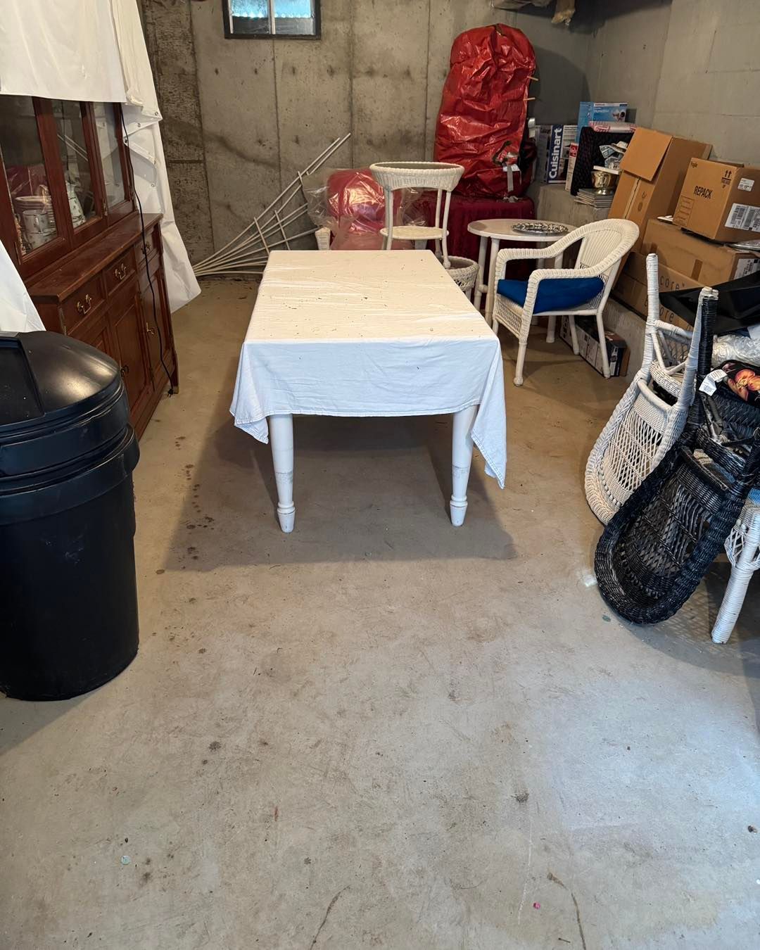 Small basement storage room with a white table, plastic chairs, boxes, and a black trash can.