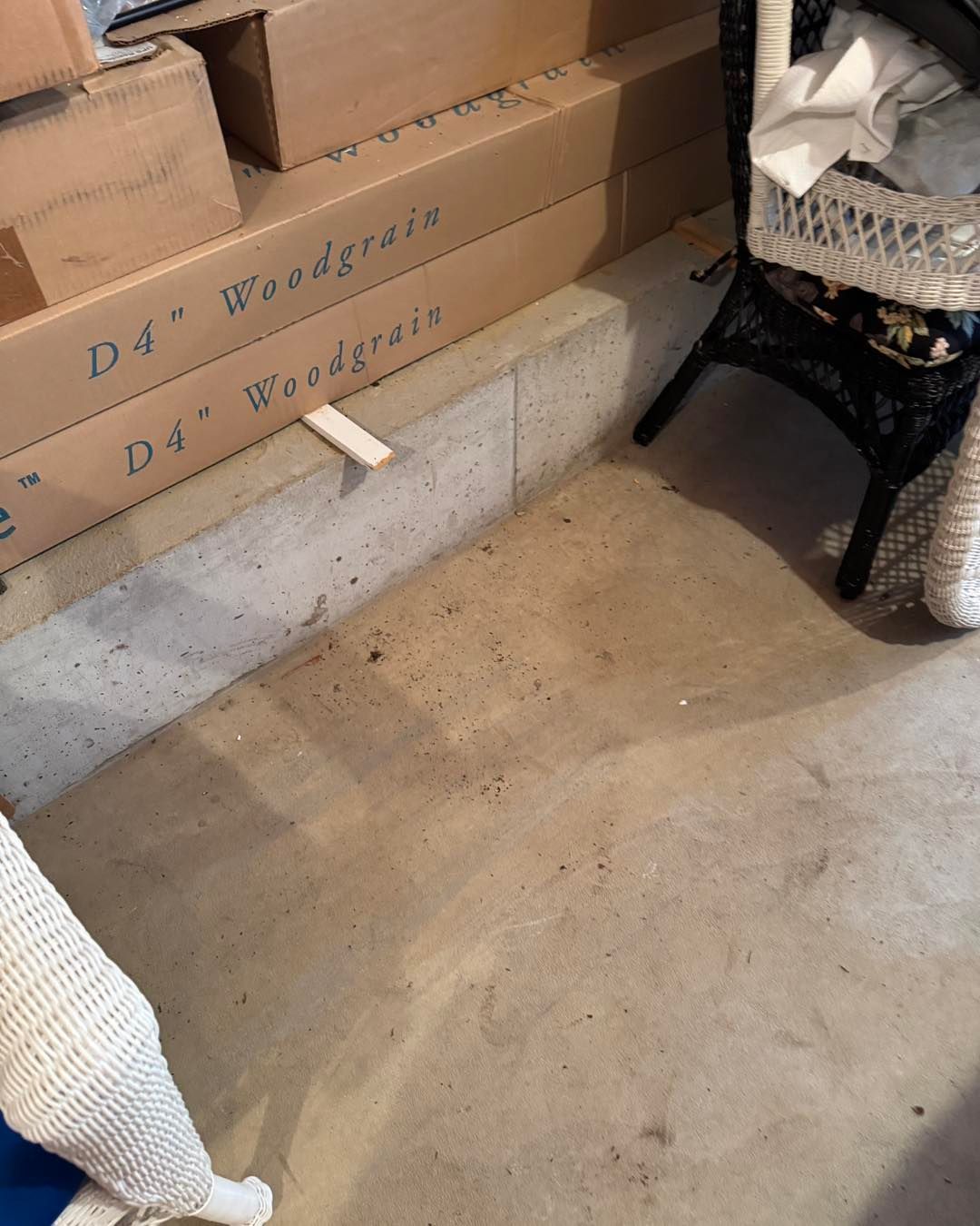 Cardboard boxes stacked on a concrete floor beside a wicker chair and a white rug in a storage area.