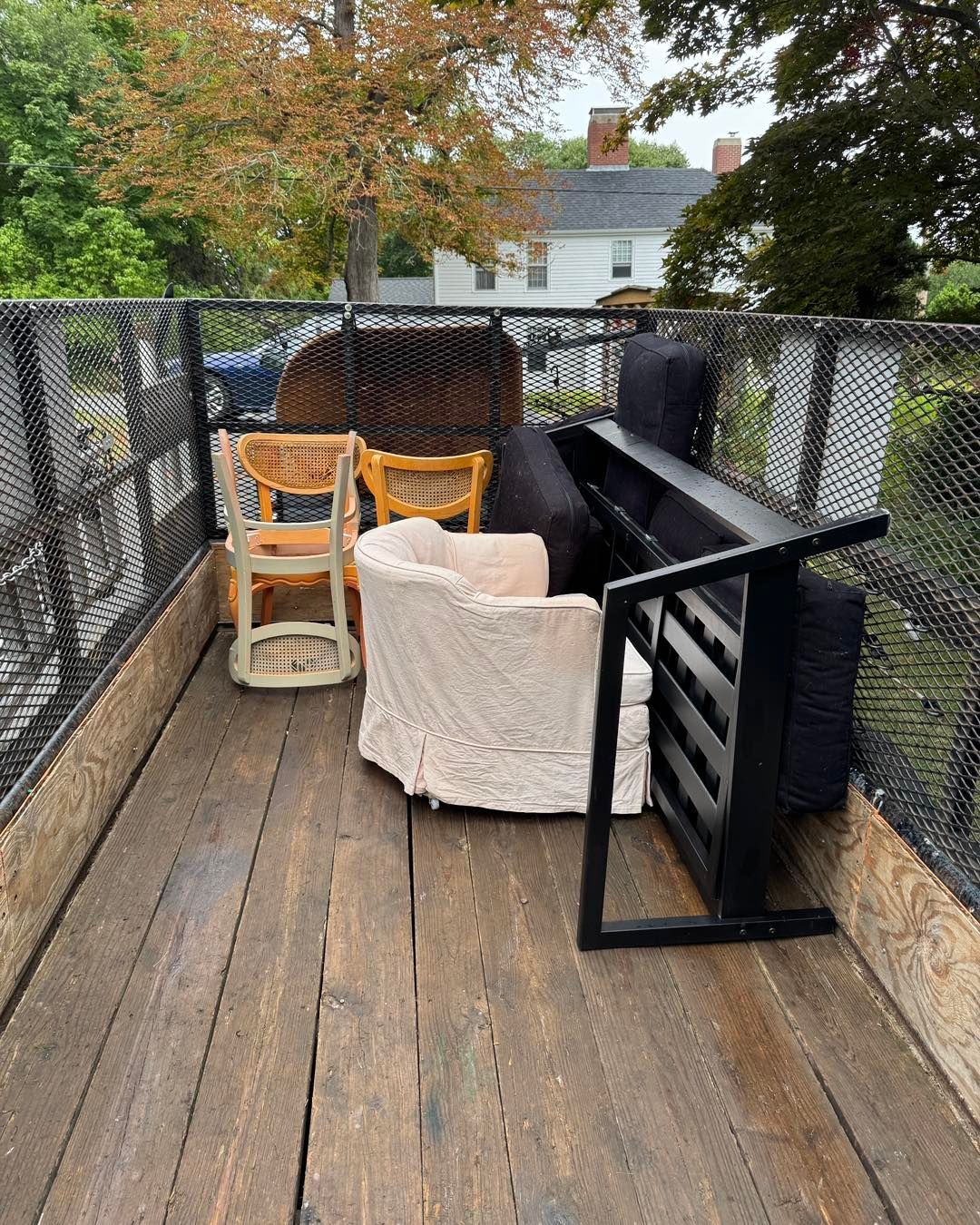 Wooden deck with black railing, wicker chairs, a round cream chair, and a grill in a backyard.