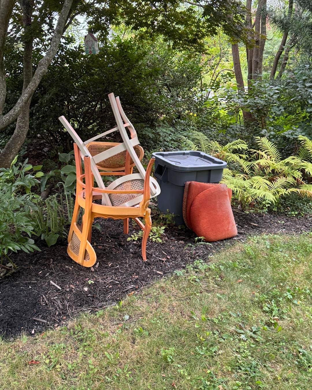 Orange chair and storage bin beside a fallen red cushion in a garden bed with trees and grass.