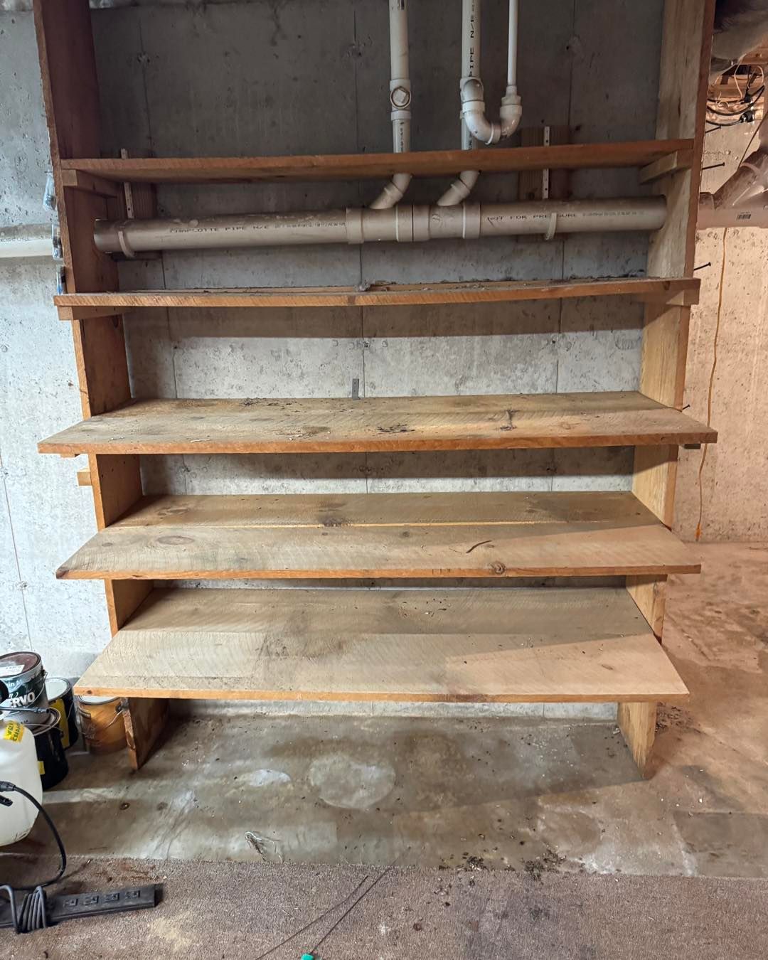 Wooden wall-mounted shelves in an unfinished basement with exposed pipes and concrete floor.