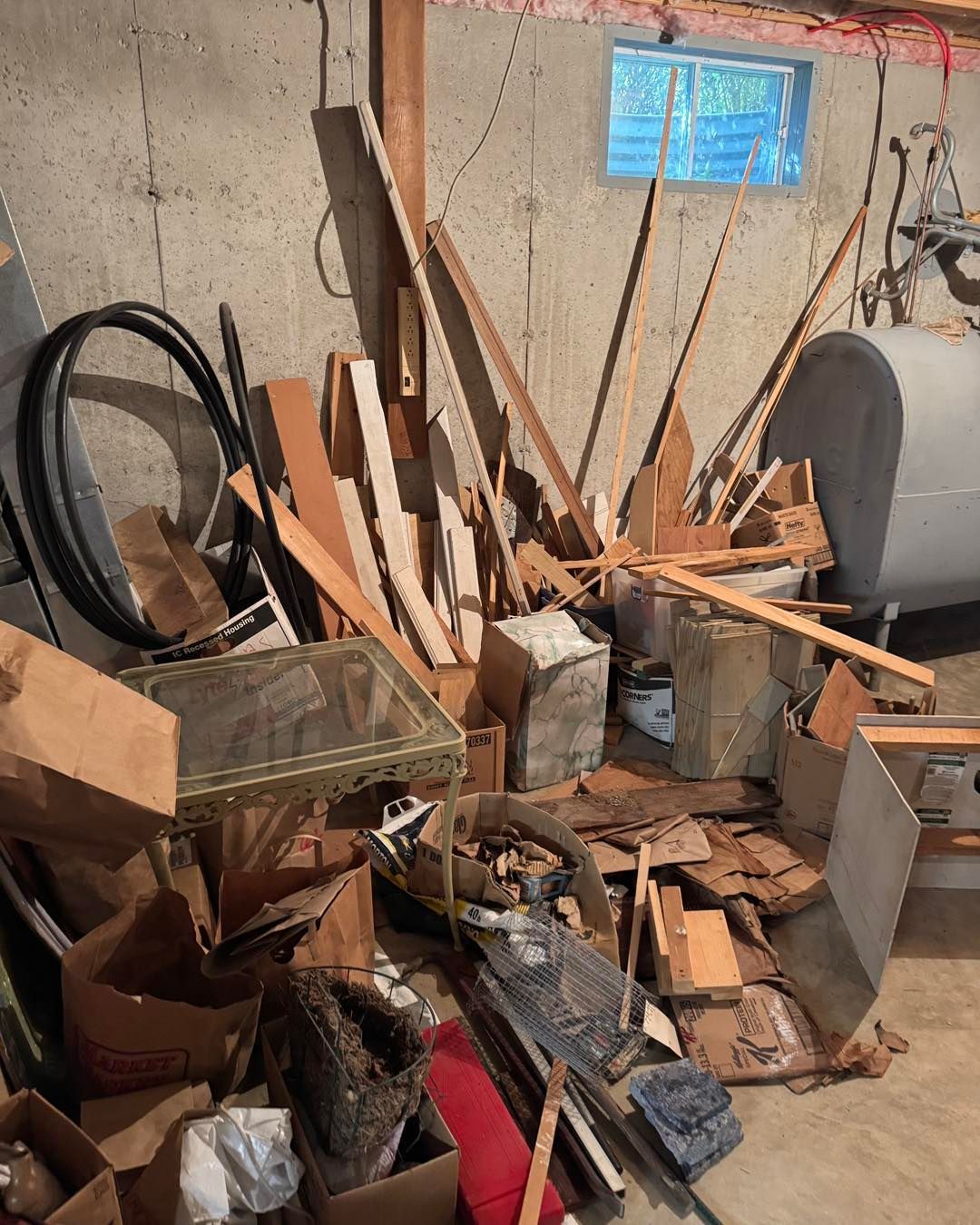 Cluttered basement with piles of wood scraps, tools, and debris against a concrete wall
