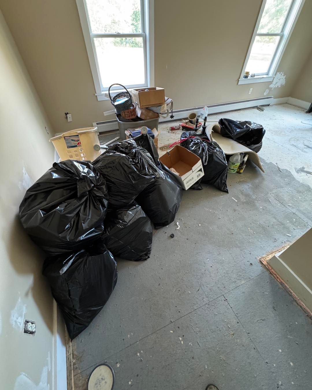 Room with scattered black trash bags, boxes, and debris on the floor near windows during cleanup.