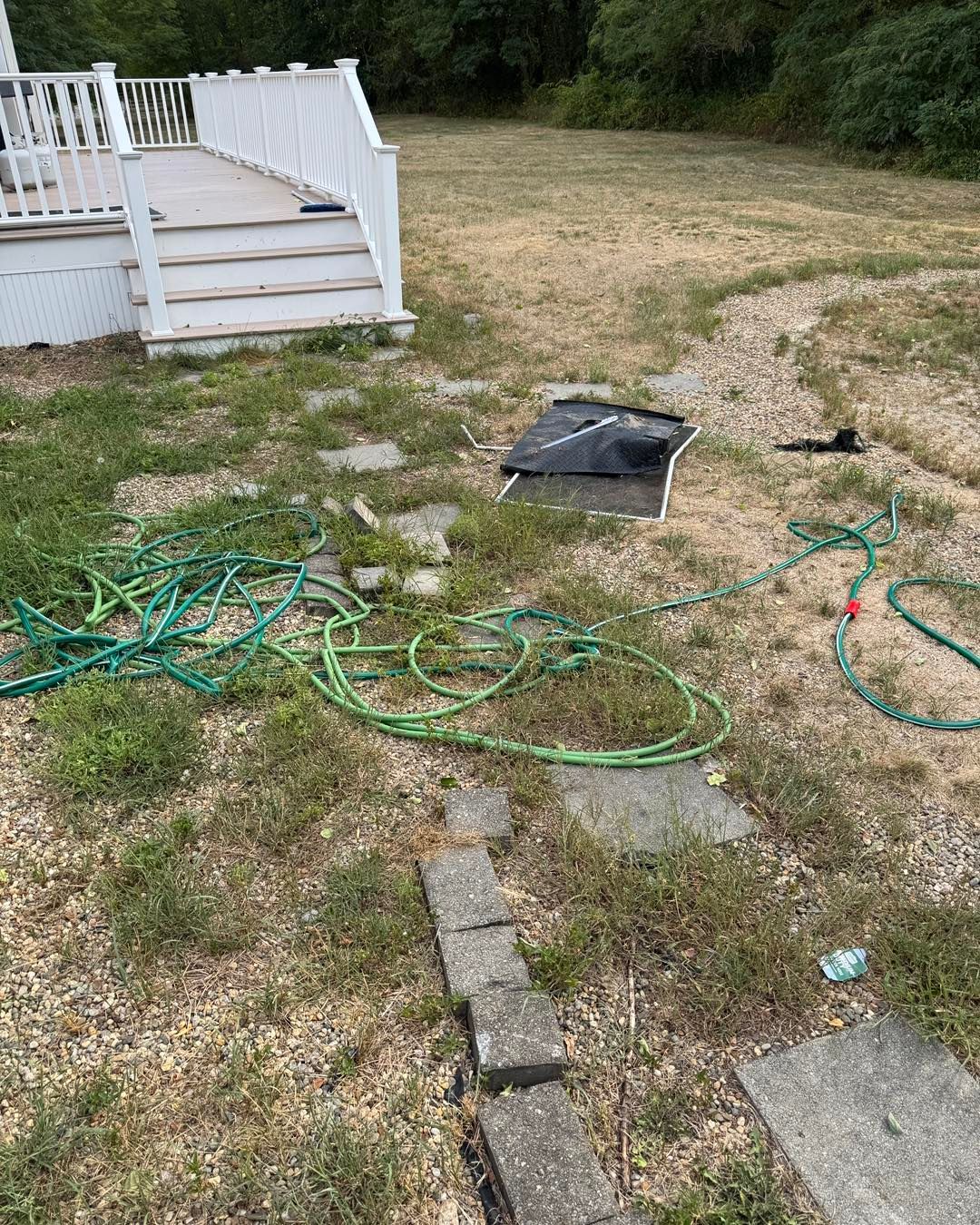 Backyard lawn with a white deck, scattered green hoses, and a fallen black frame on dry grass.