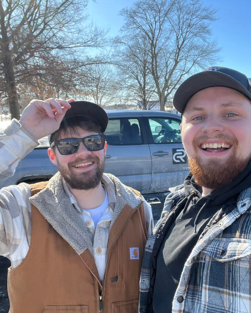Two smiling friends posing outdoors in winter, one tipping his cap beside a parked truck.