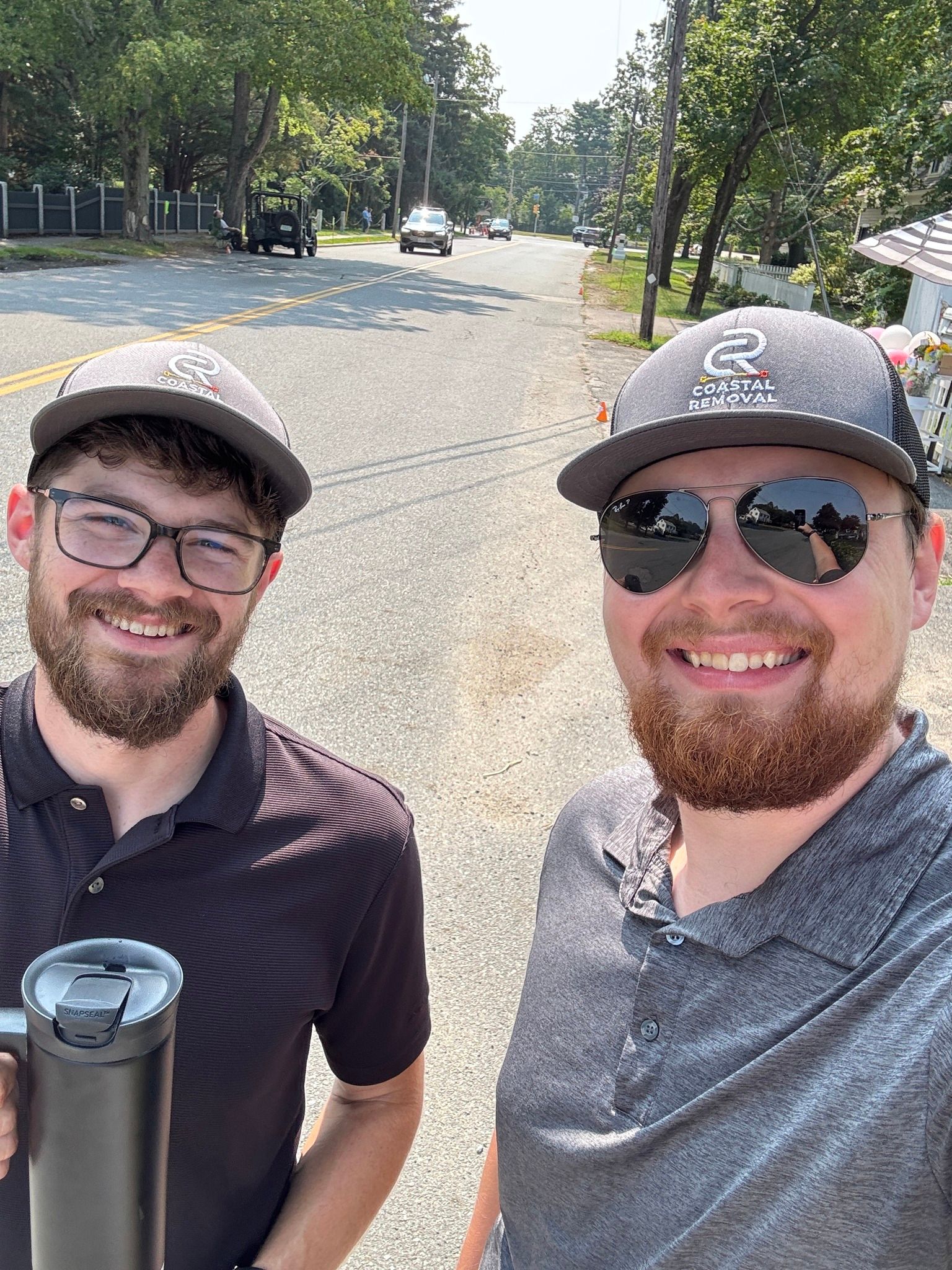 Two smiling men wearing caps and sunglasses standing on a tree-lined street, one holding a selfie stick