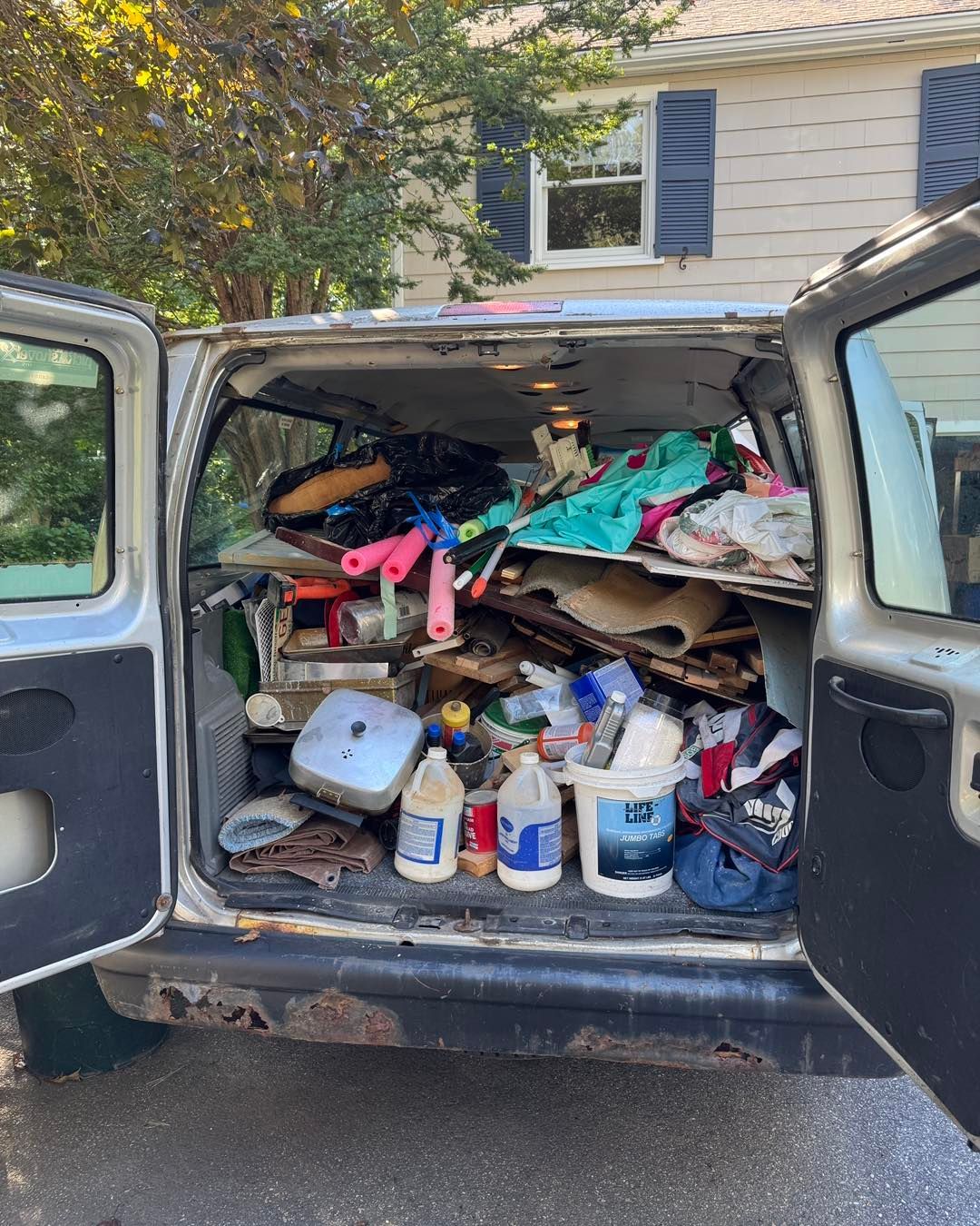 Open car trunk packed with clutter, including bags, wood scraps, and plastic bottles, outside a house