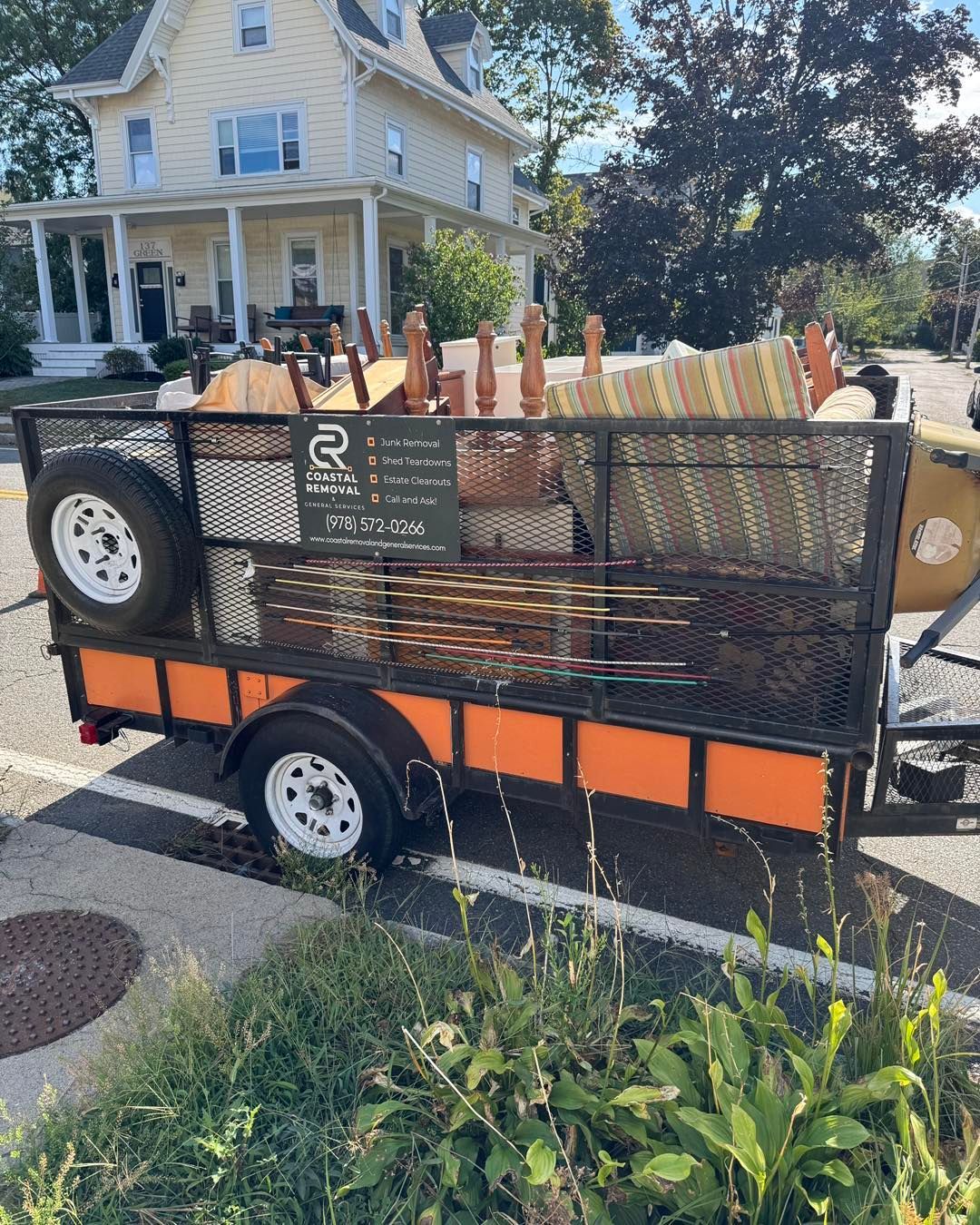 Trailer with orange panels and mesh sides parked on a street in front of a house and trees