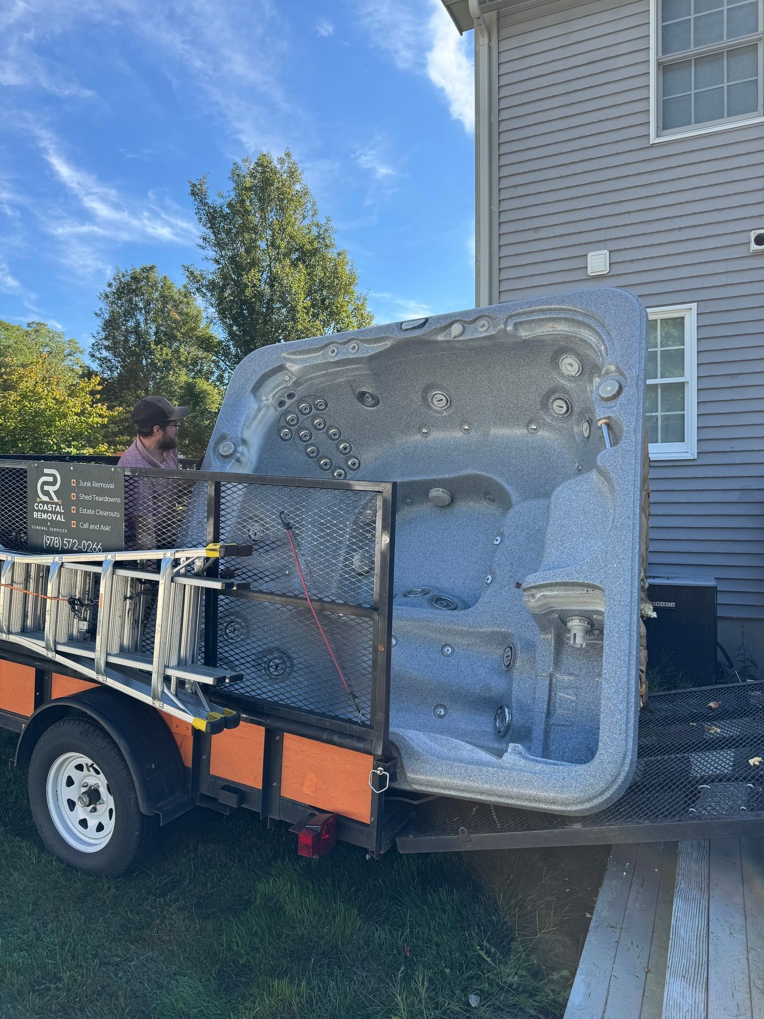 Large gray industrial machine on a flatbed trailer beside a house, with a person securing it.