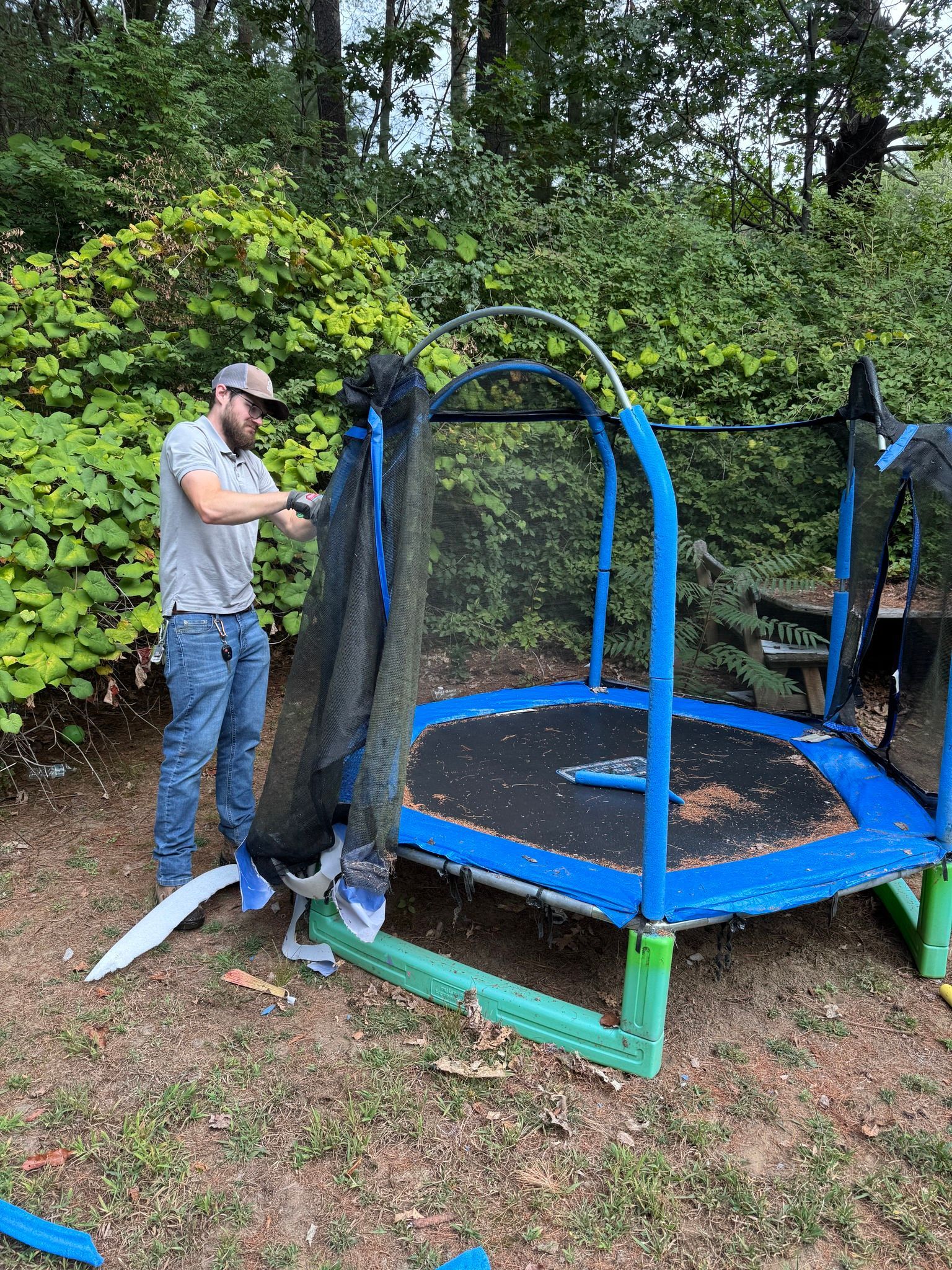 Man setting up a blue-and-black trampoline in a grassy yard with trees behind it