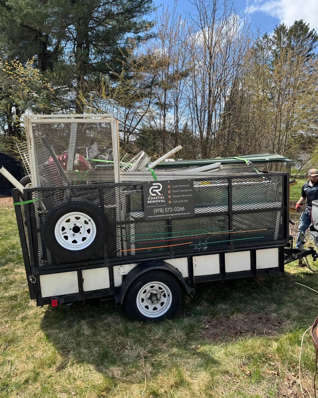 Small black utility trailer with mesh sides and white wheels parked on grass in a wooded yard.