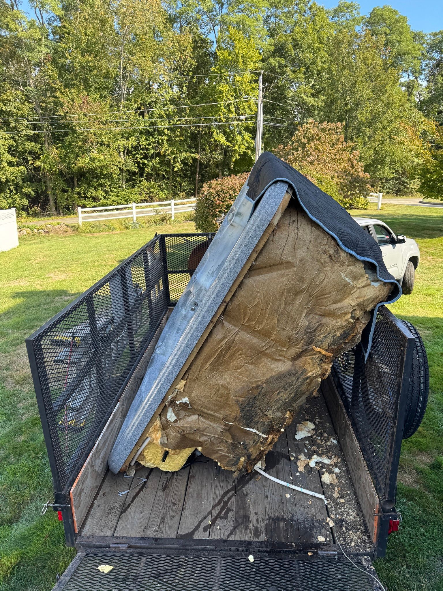 Large broken mattress and metal frame piled in a black utility trailer outdoors