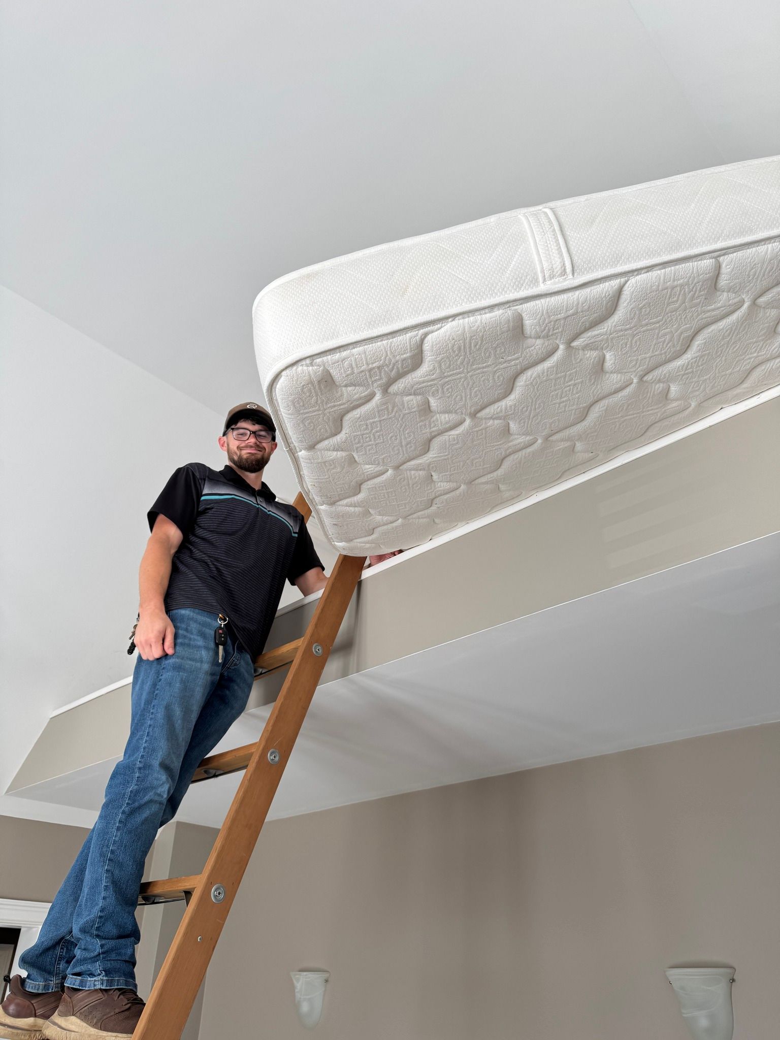Man standing on a ladder beneath a mattress, inspecting or moving it near a ceiling