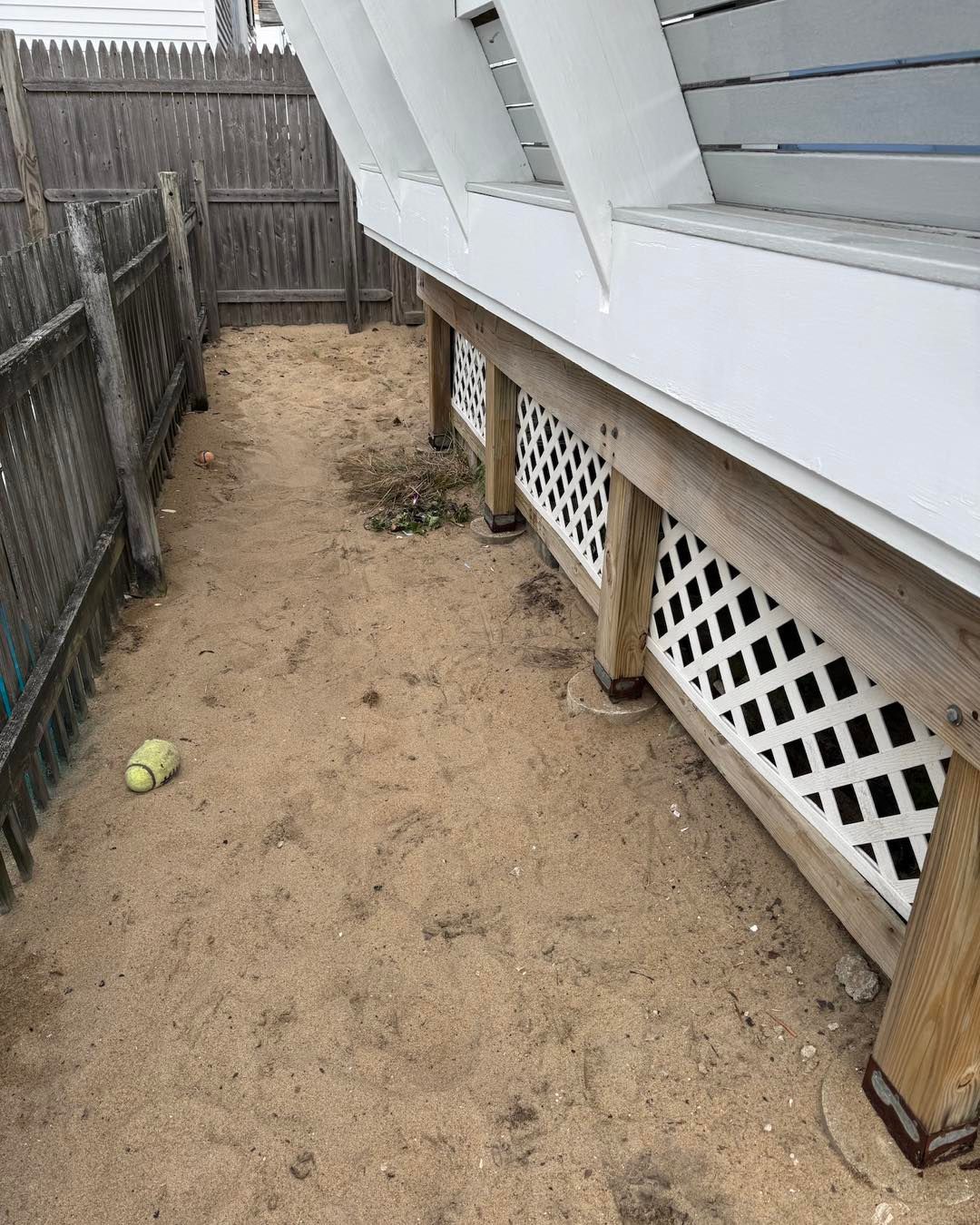 Narrow sandy side yard beside a house with lattice skirting, fence, and a tennis ball on the ground