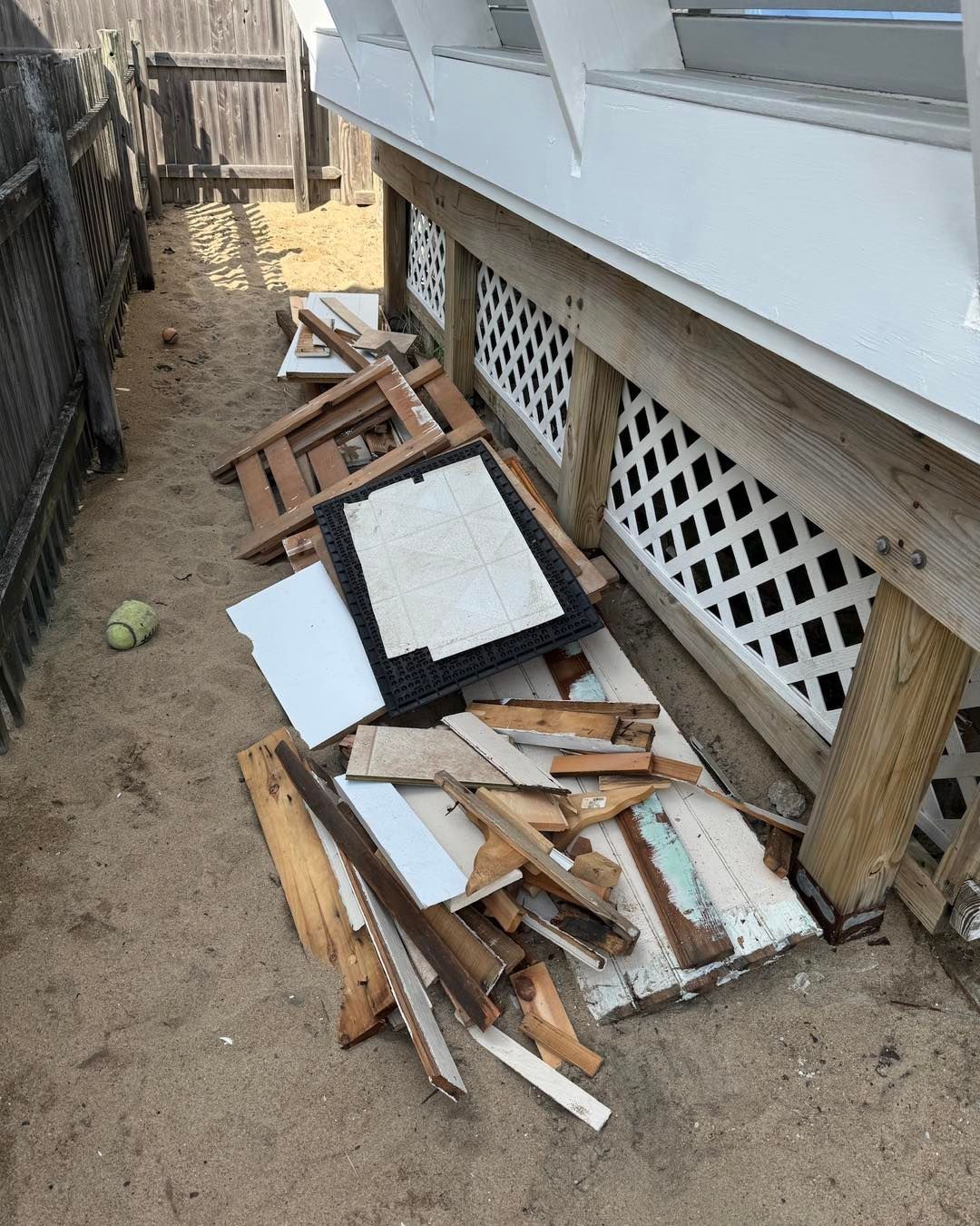 Pile of broken white and brown wood debris beside a house foundation in a narrow dirt side yard.