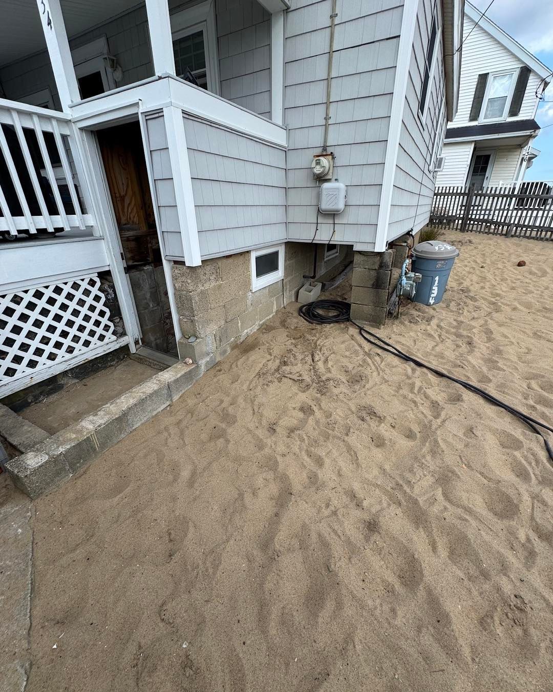 Sand beside a gray house with a deck, basement window, utility meter, and hose line.