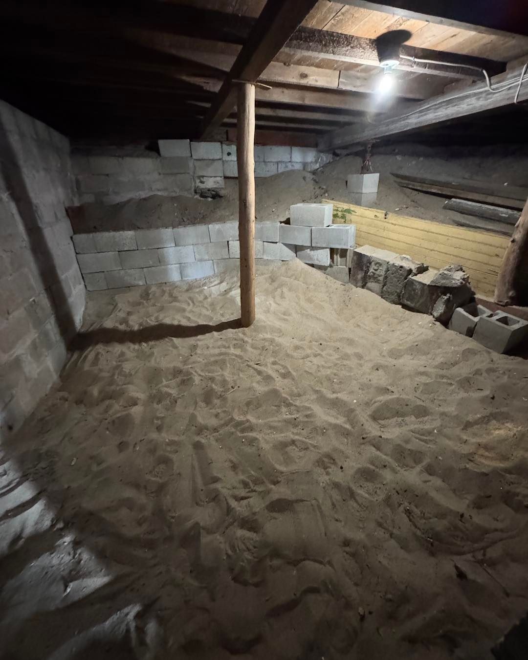 Dim basement crawlspace with sandy floor, cinder block walls, and a single hanging light bulb