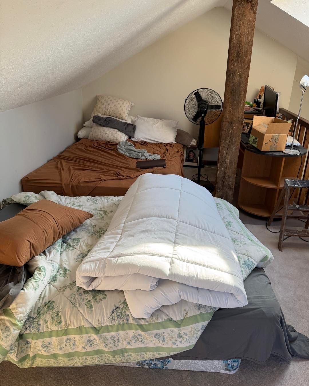 Small attic bedroom with two unmade beds, pillows, blankets, and a fan beside a wooden post.