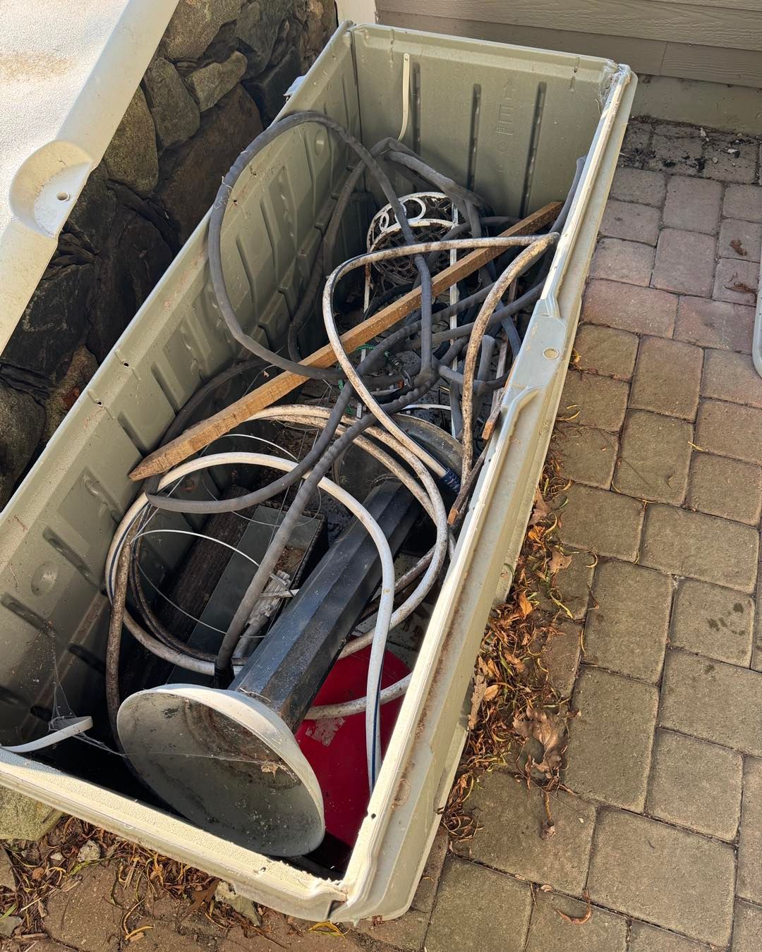 A plastic bin filled with tangled cables, hoses, and metal parts on a brick patio.